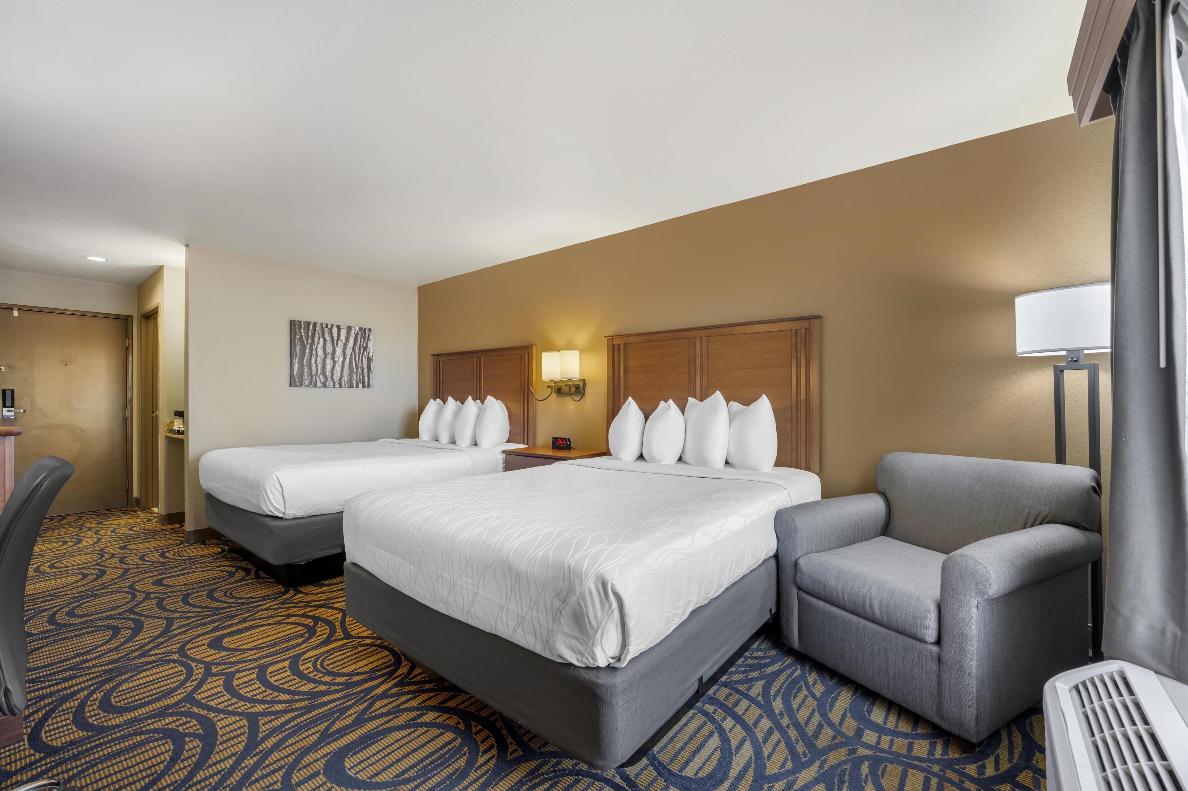 A hotel room with two beds, a desk, a chair, and a lamp.