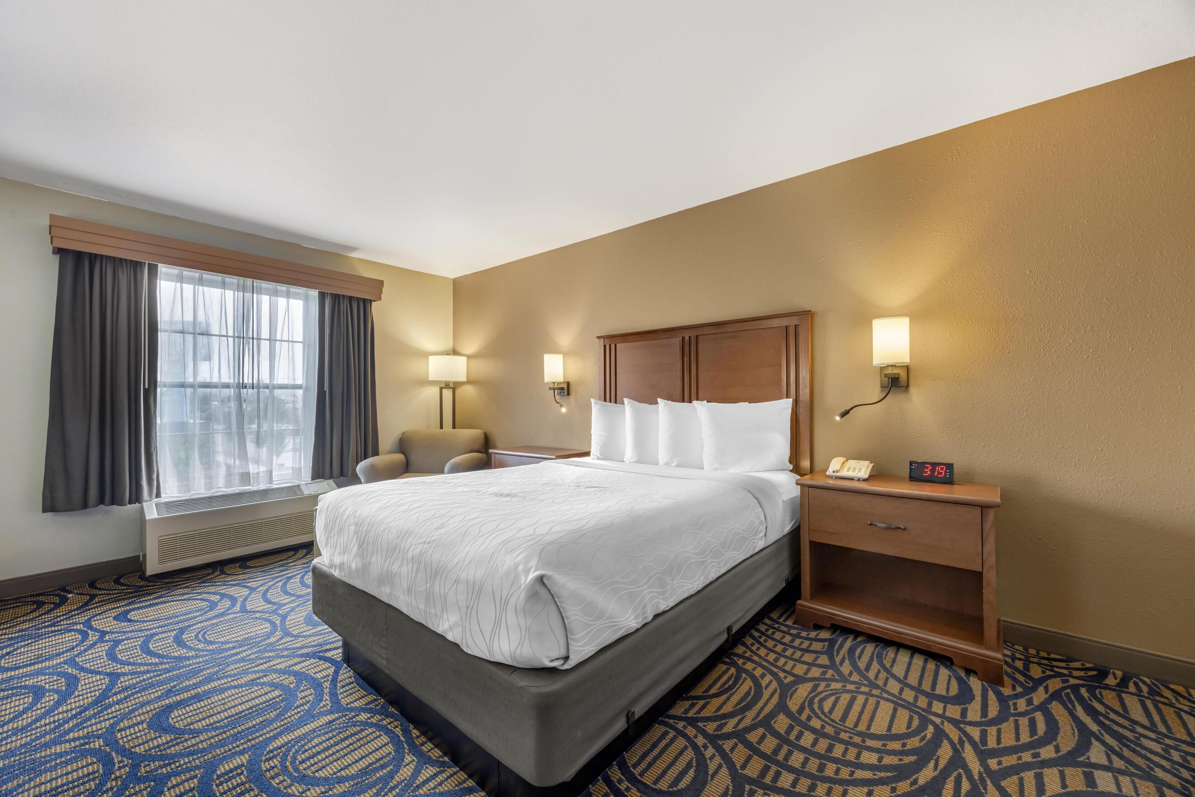 A hotel room with a large bed, two bedside lamps, a nightstand, a chair, and a window with curtains.