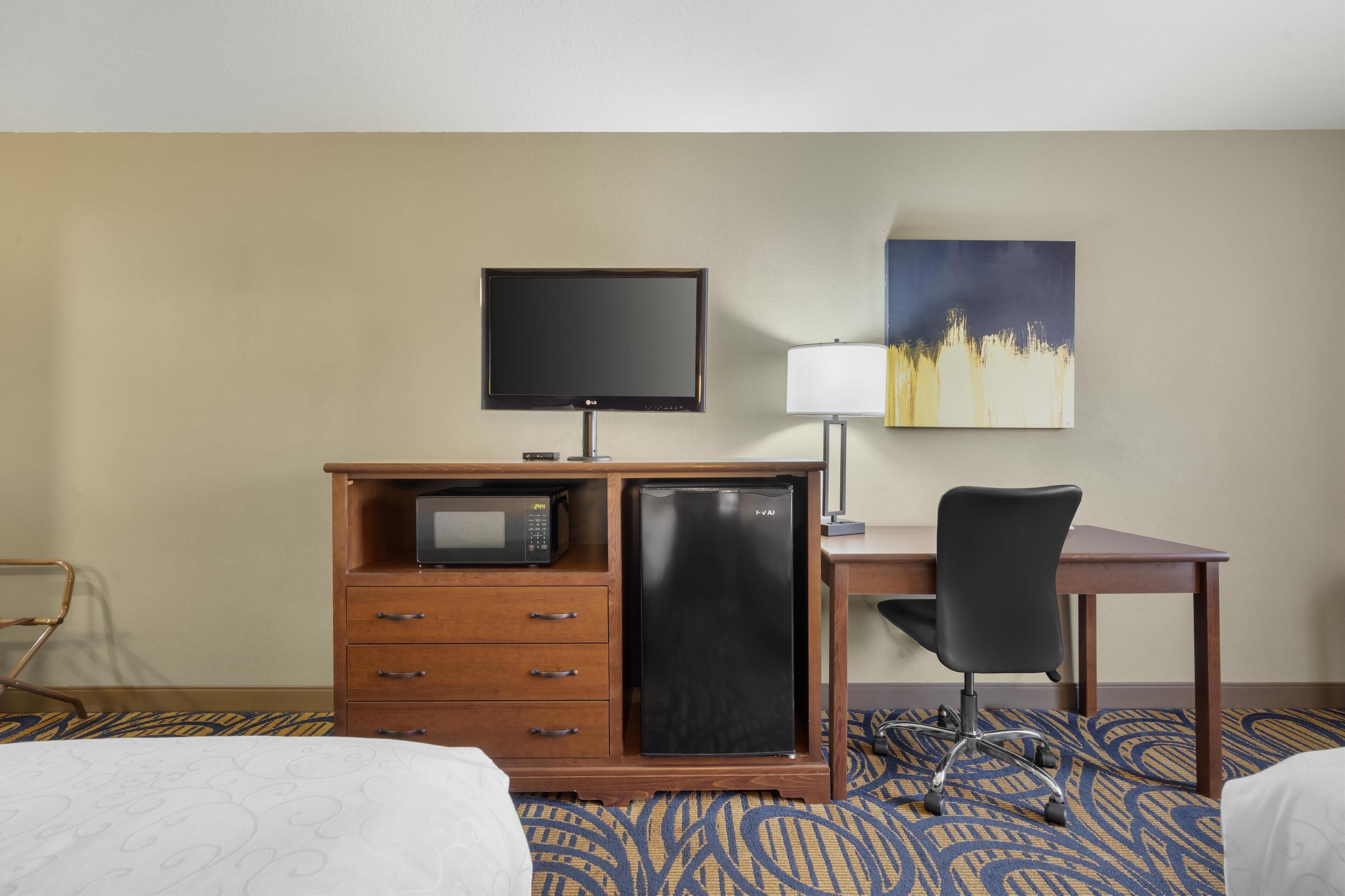 Ein Hotelzimmer mit einem Bett, einem Schreibtisch mit Computer, einem Fernseher, einem Kühlschrank, einer Mikrowelle, einer Lampe und einem Stuhl.