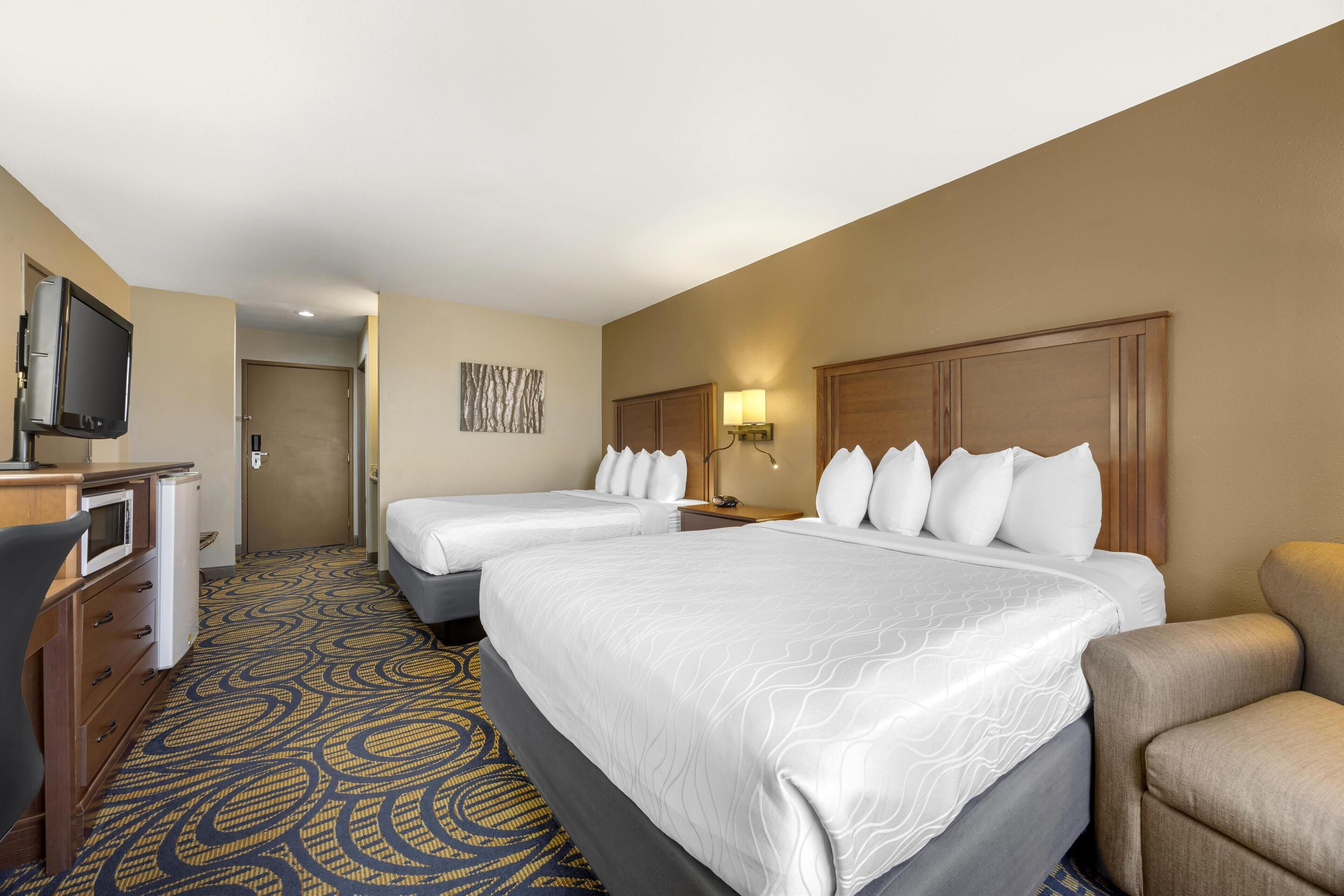 A hotel room with two beds, a desk with a television, and a patterned carpet.