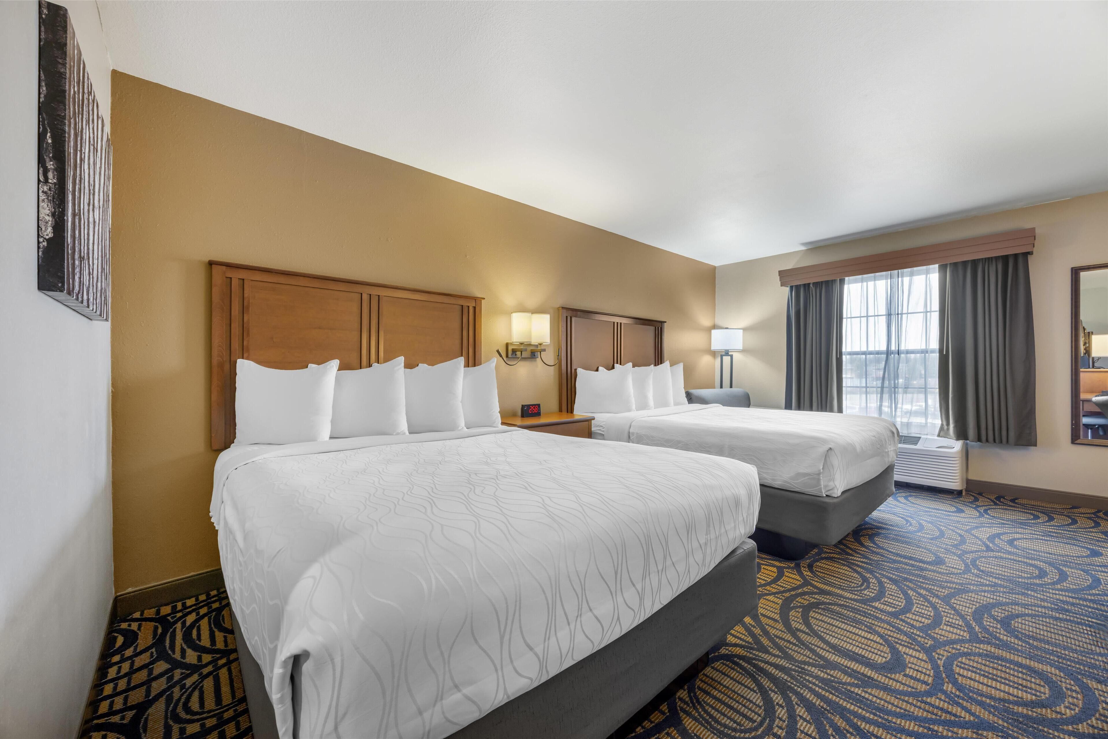 A hotel room with two beds, a large window with curtains, a wall-mounted clock, and a patterned carpet.