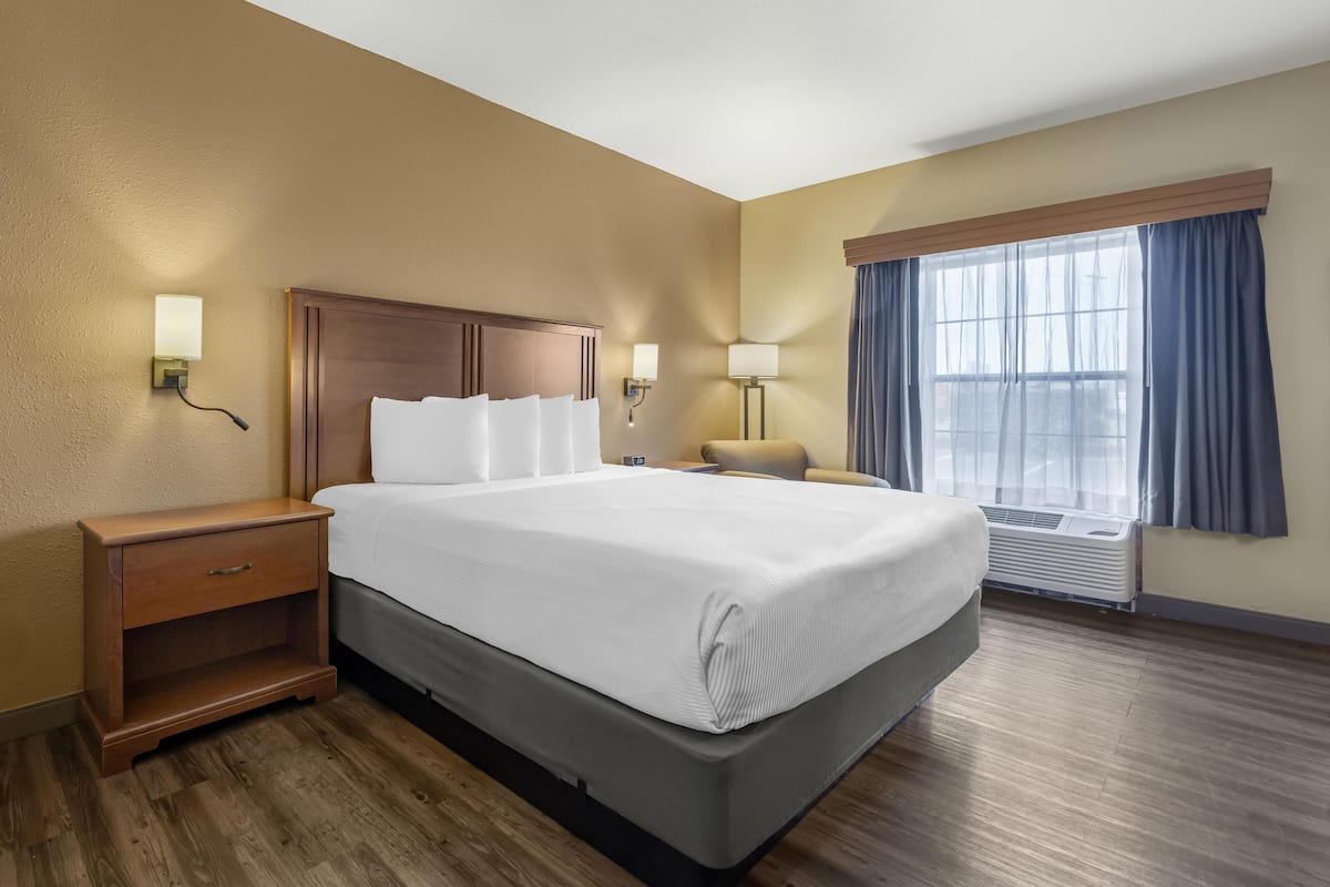 Habitación de hotel con una cama grande, mesita de noche de madera, dos lámparas fijadas a la pared, una ventana con cortinas y un equipo de aire acondicionado.