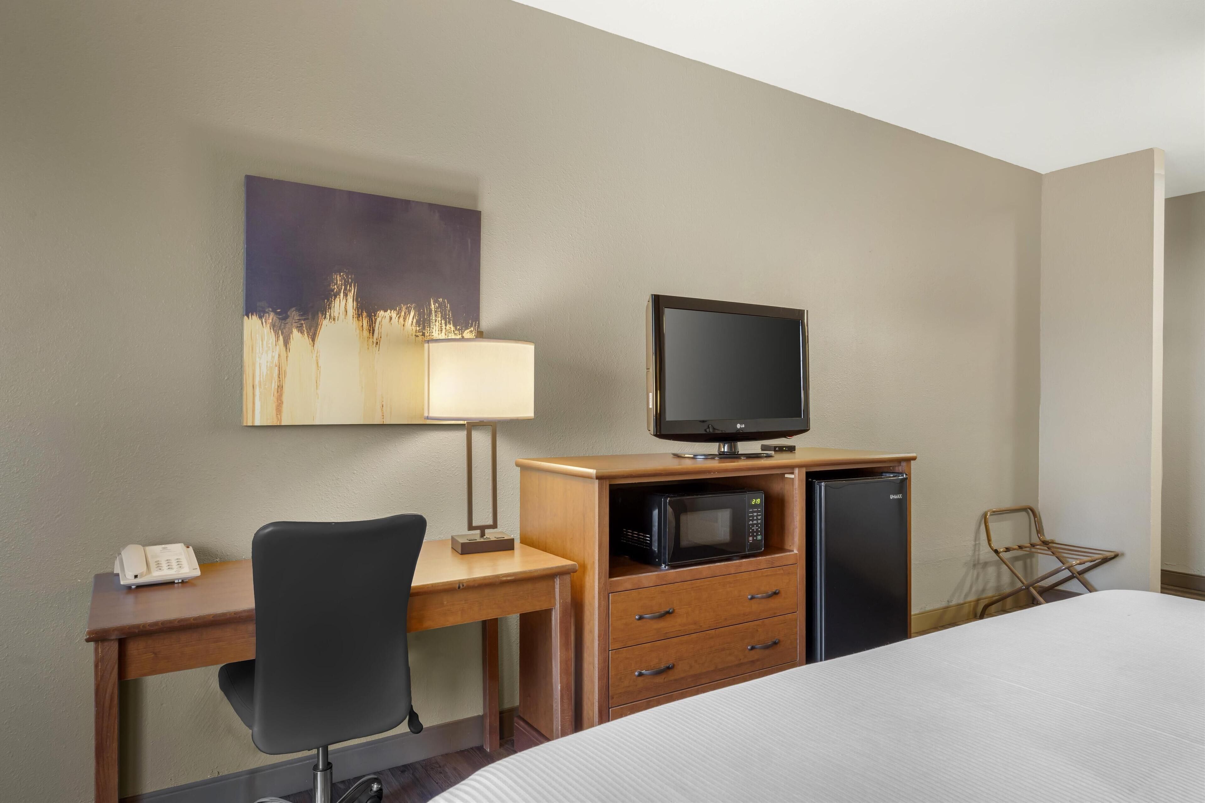 A hotel room with a bed, a desk with a computer, a television, a microwave, a telephone, and a painting on the wall.