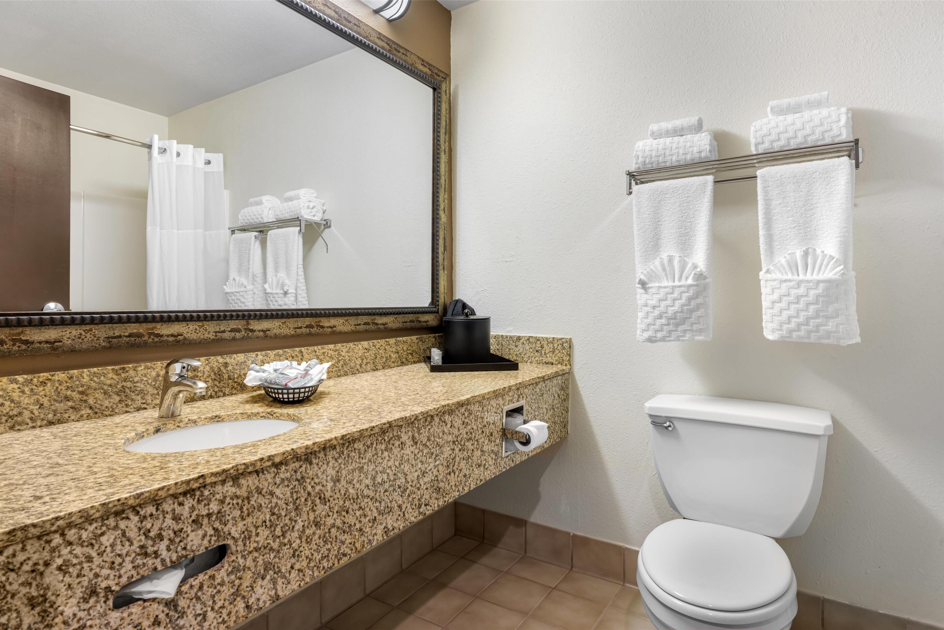 A bathroom with a granite countertop sink, a mirror, a towel rack with towels, and a white toilet.