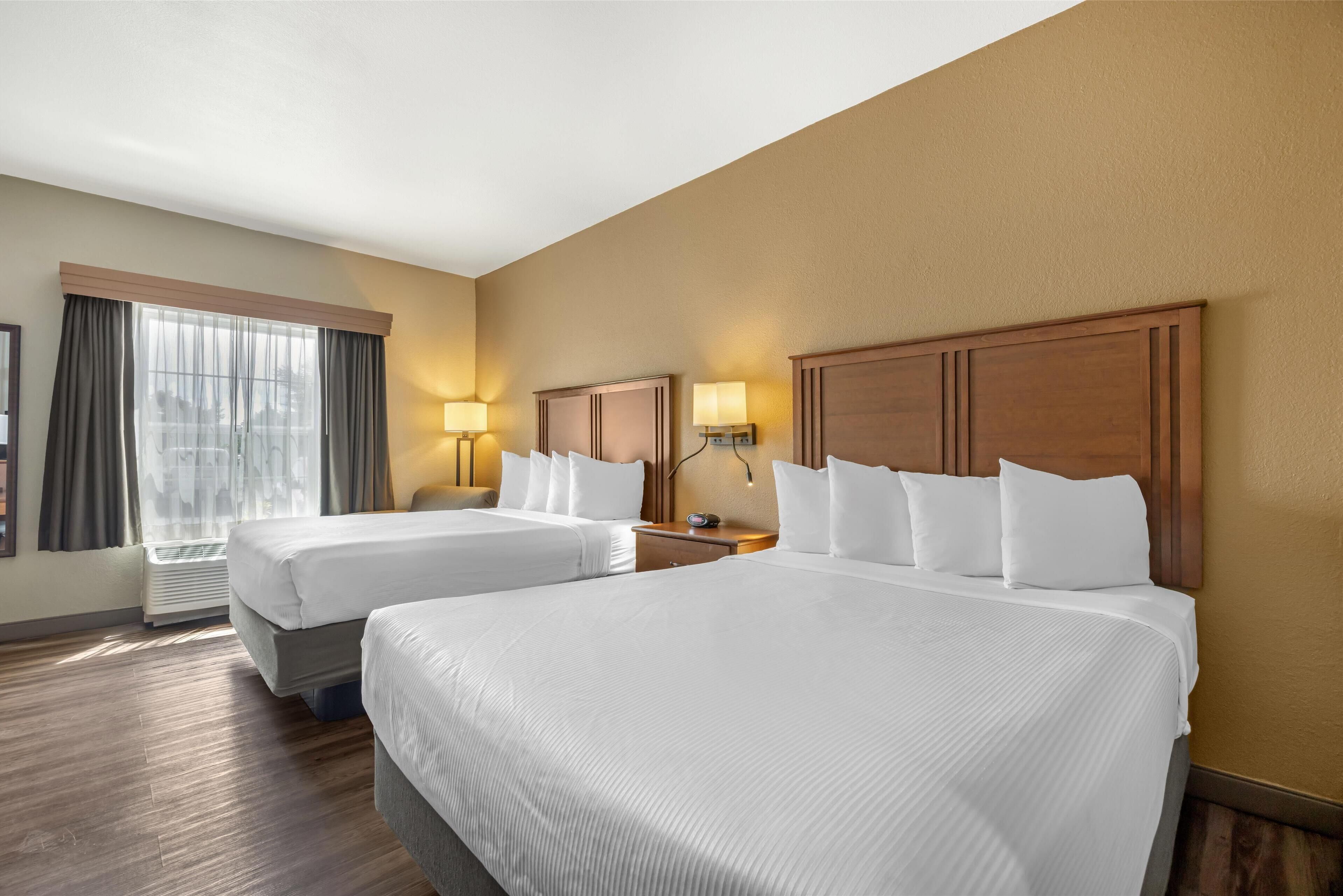 A hotel room with two beds, a large window with curtains, a wall-mounted heater, and two bedside lamps.