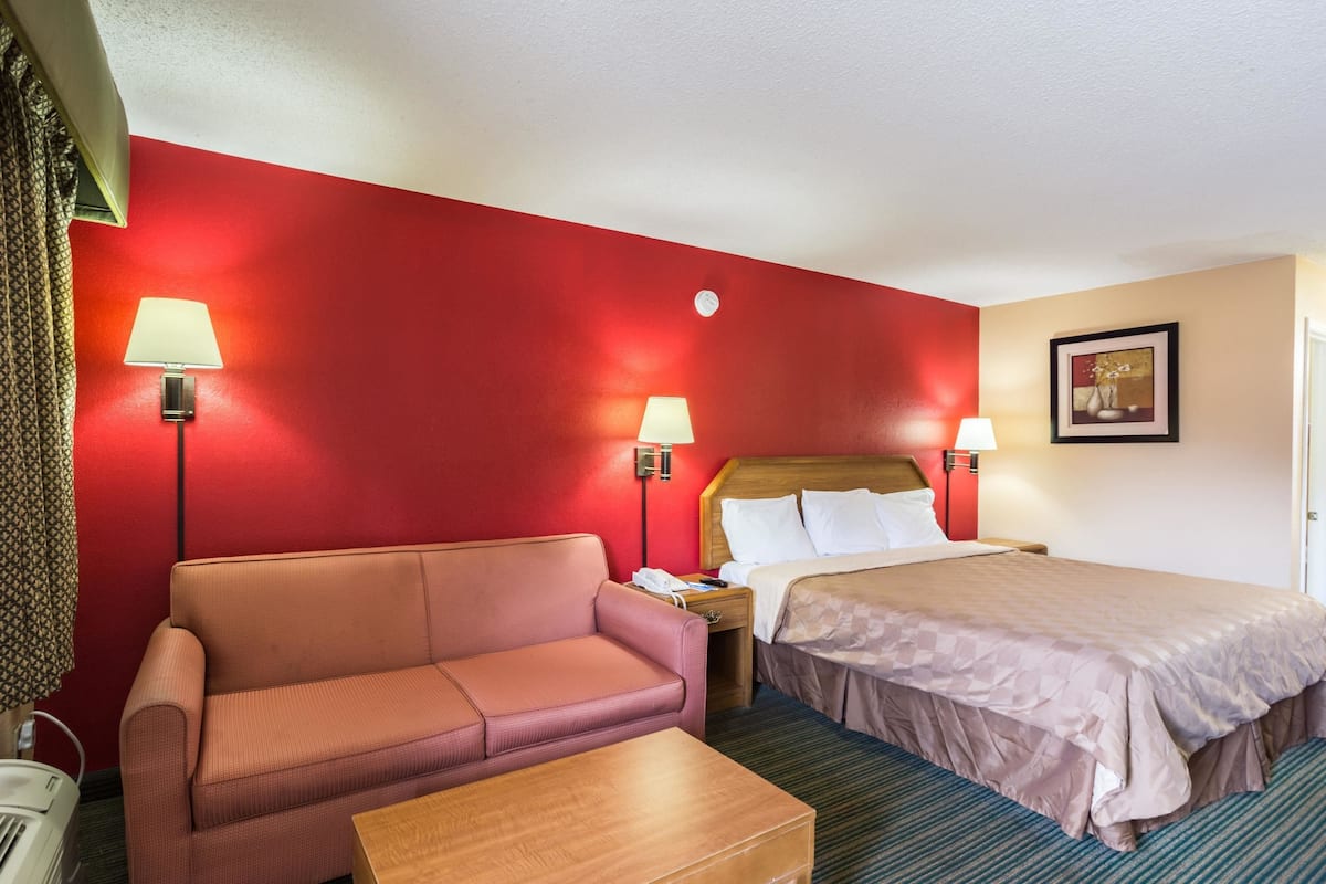 A hotel room with a red accent wall, a sofa, a bed, a nightstand, a lamp, and a framed picture on the wall.