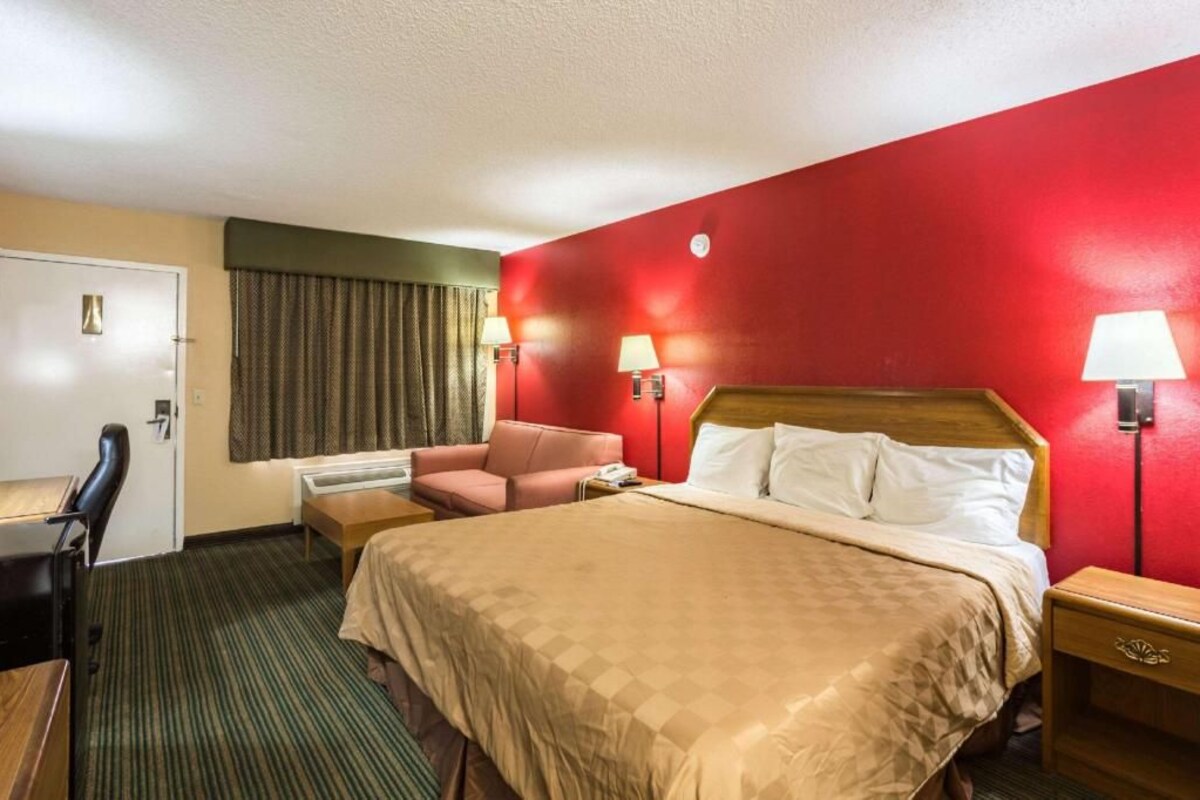 Una habitación de hotel con cama, escritorio, silla, lámpara de escritorio y mesita de noche.