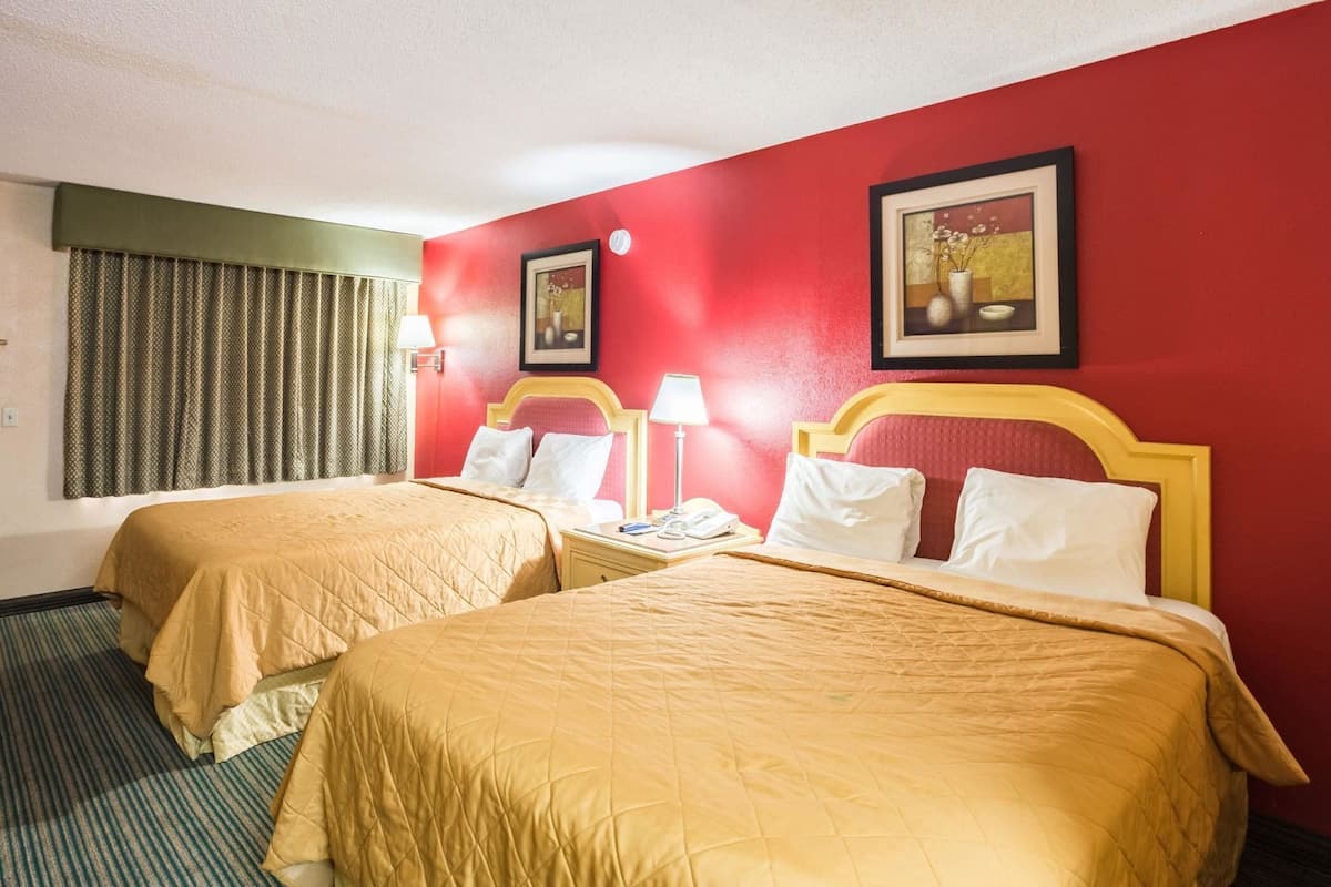 A hotel room with two beds, a window with curtains, a framed picture on the wall, and a nightstand with a lamp.