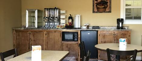 A small kitchenette with a coffee machine, microwave, and refrigerator. There are two tables and four chairs with plaid cushions. A sign on the wall reads "Community Coffee."