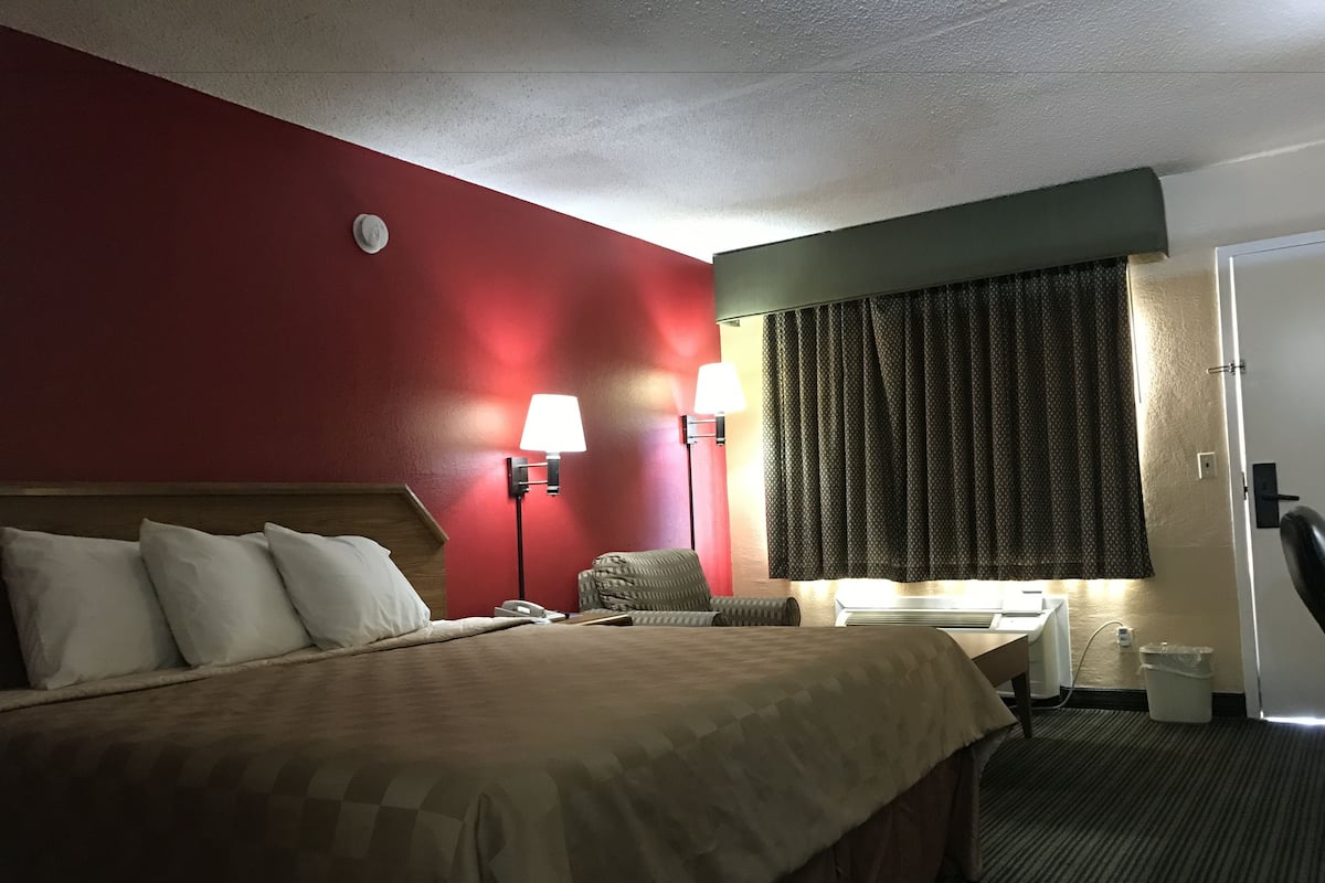 A hotel room with a large bed, two bedside lamps, a desk with a chair, a telephone, and a window with curtains.