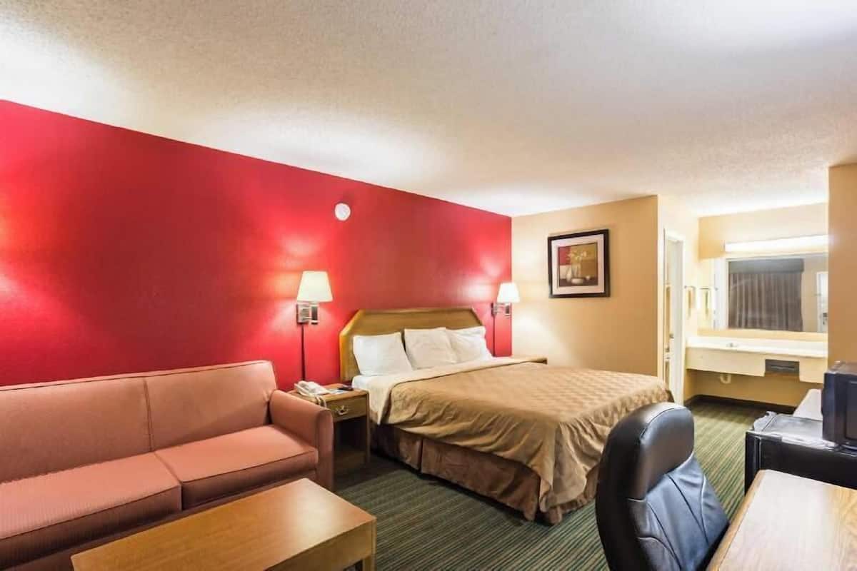 A hotel room with a red accent wall, a bed with a beige bedspread, a wooden nightstand, a desk with a chair, and a window with curtains.