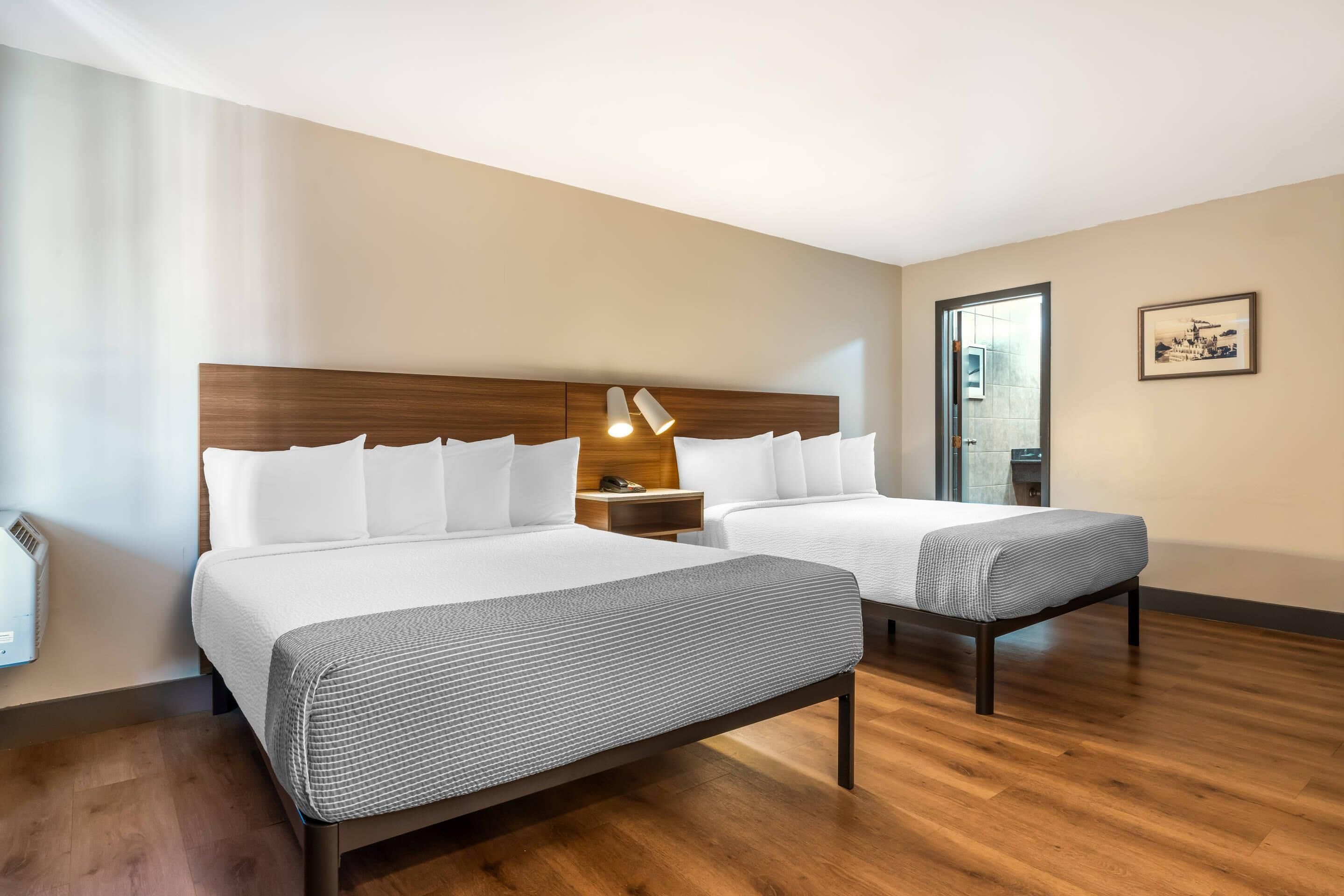 Quarto de hotel com duas camas, cabeceira de madeira, mesa de cabeceira com abajur e um quadro na parede.