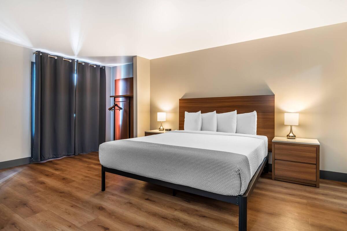 Une chambre d’hôtel moderne avec un grand lit, une tête de lit en bois, deux tables de chevet avec des lampes, une armoire et un plancher en bois.