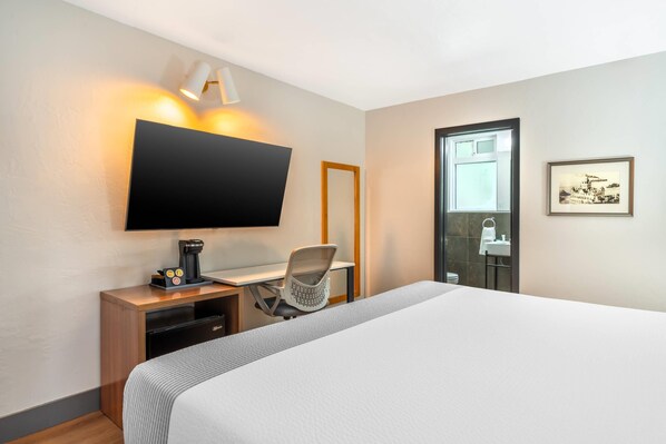 Une chambre d’hôtel comprenant un lit, un bureau avec une machine à café, une chaise, un téléviseur fixé au mur et une salle de bain avec un lavabo et un tableau encadré.