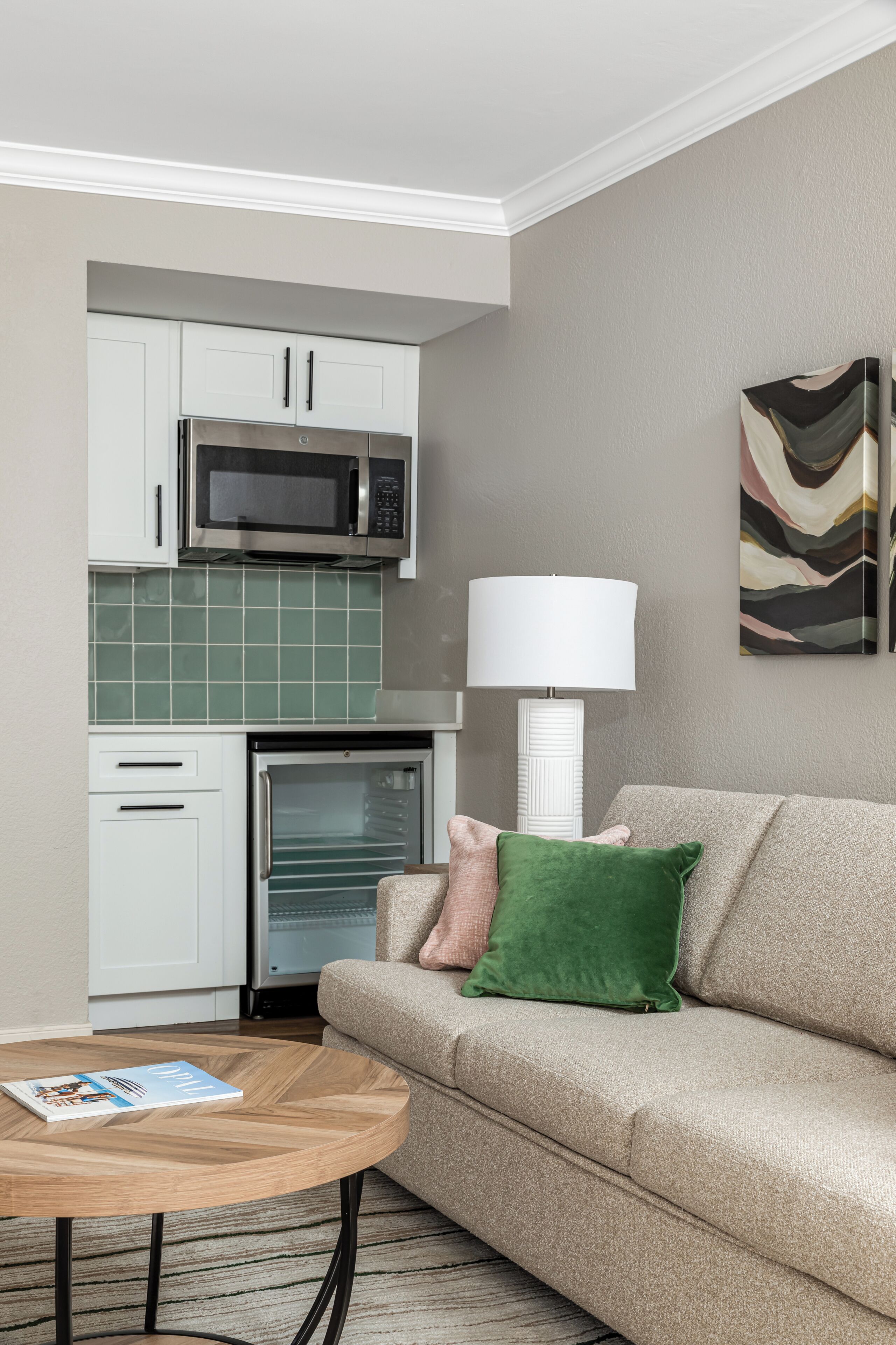 Una sala moderna con un sofá color beige, una mesita de centro, una revista, un microondas, un refrigerador y un cuadro en la pared.