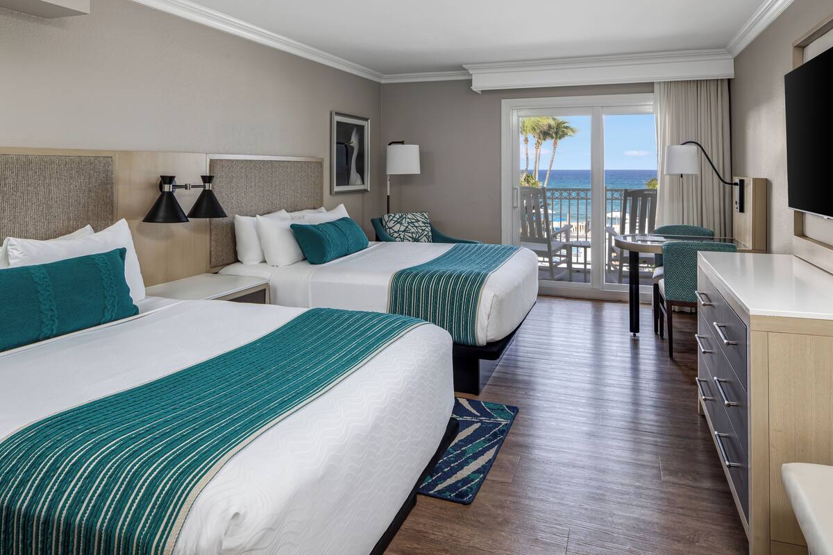 A hotel room with two beds, a desk, a chair, a TV, and a balcony with a view of the ocean.