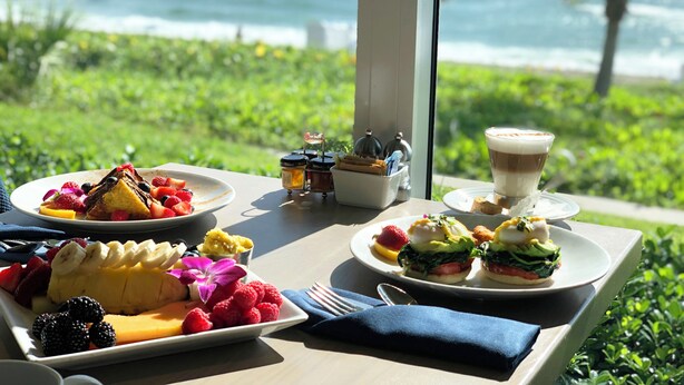 A table with various breakfast dishes, a coffee cup, and condiments, set against a backdrop of a lush green landscape and the ocean. - Delray Sands Resort (Highland Beach)