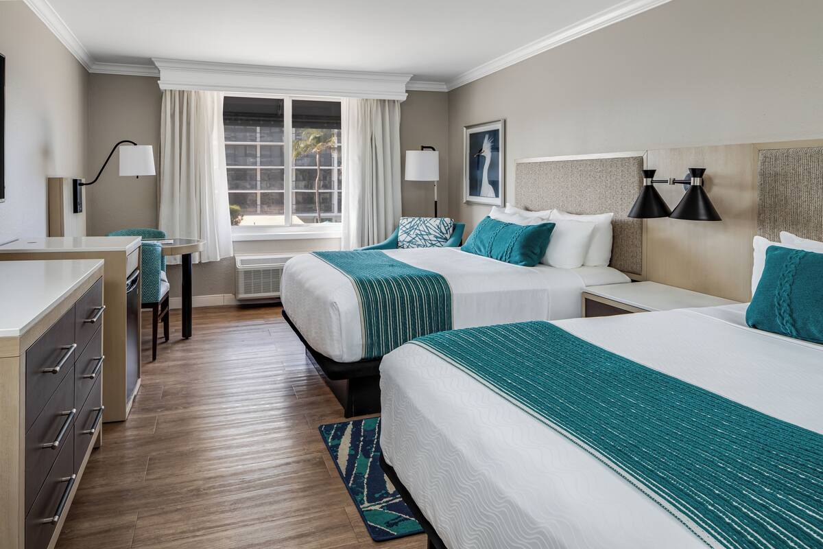 A hotel room with two beds, a desk, a chair, a window with curtains, and a wall-mounted lamp.