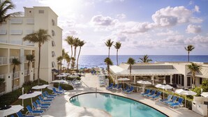 A pool area with lounge chairs and umbrellas, a curved pool, and a view of the ocean. - Delray Sands Resort (Highland Beach)