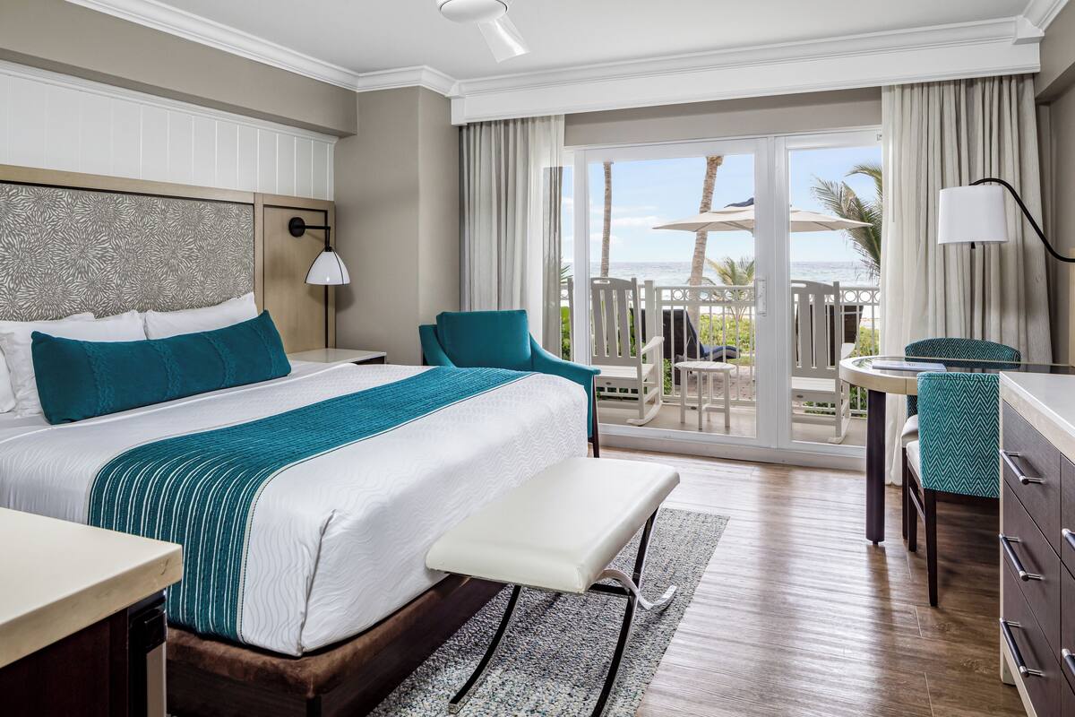 A hotel room with a large bed, a desk, a chair, and a balcony with a view of the ocean.