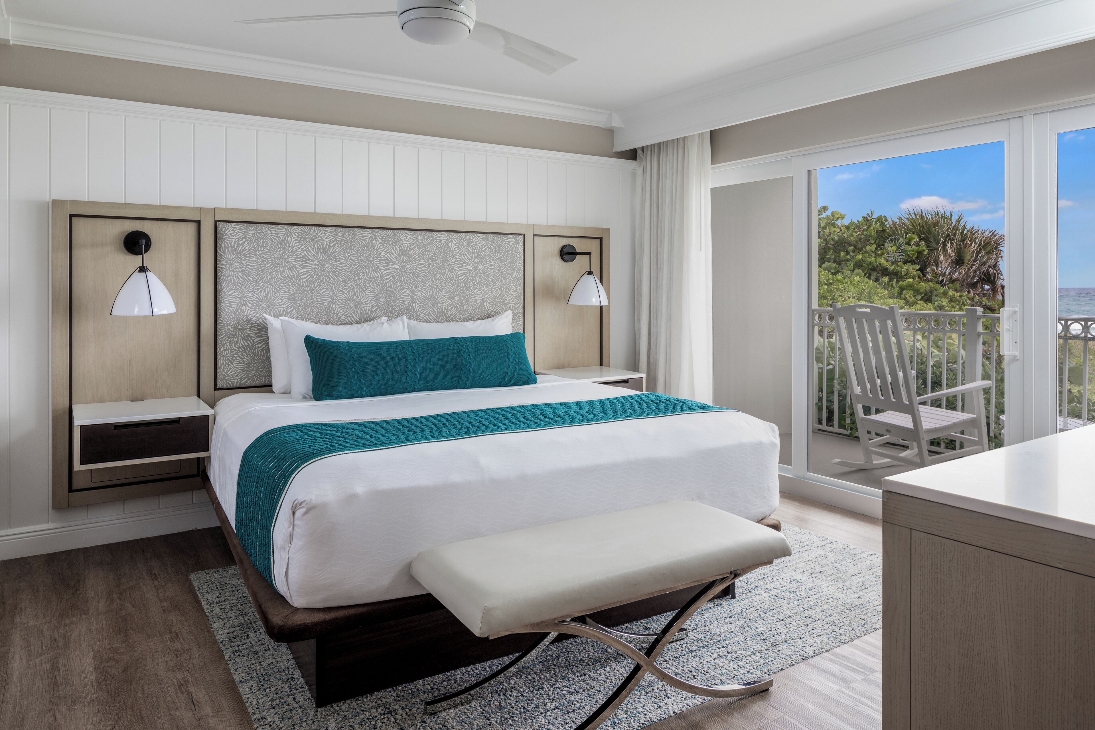 A modern hotel room with a large bed, a bench, a desk, and a balcony with a view of the ocean.