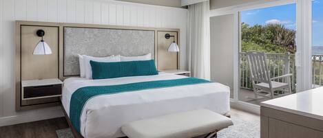 A modern hotel room with a large bed, a bench, a desk, and a balcony with a view of the ocean.