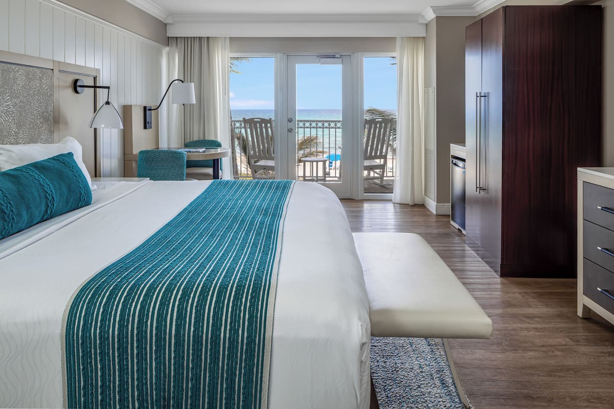 A hotel room with a large bed, a bench, a desk, and a view of the ocean through the balcony doors.