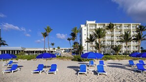 A beach with blue lounge chairs and umbrellas, a hotel building with balconies, and palm trees. - Delray Sands Resort (Highland Beach)