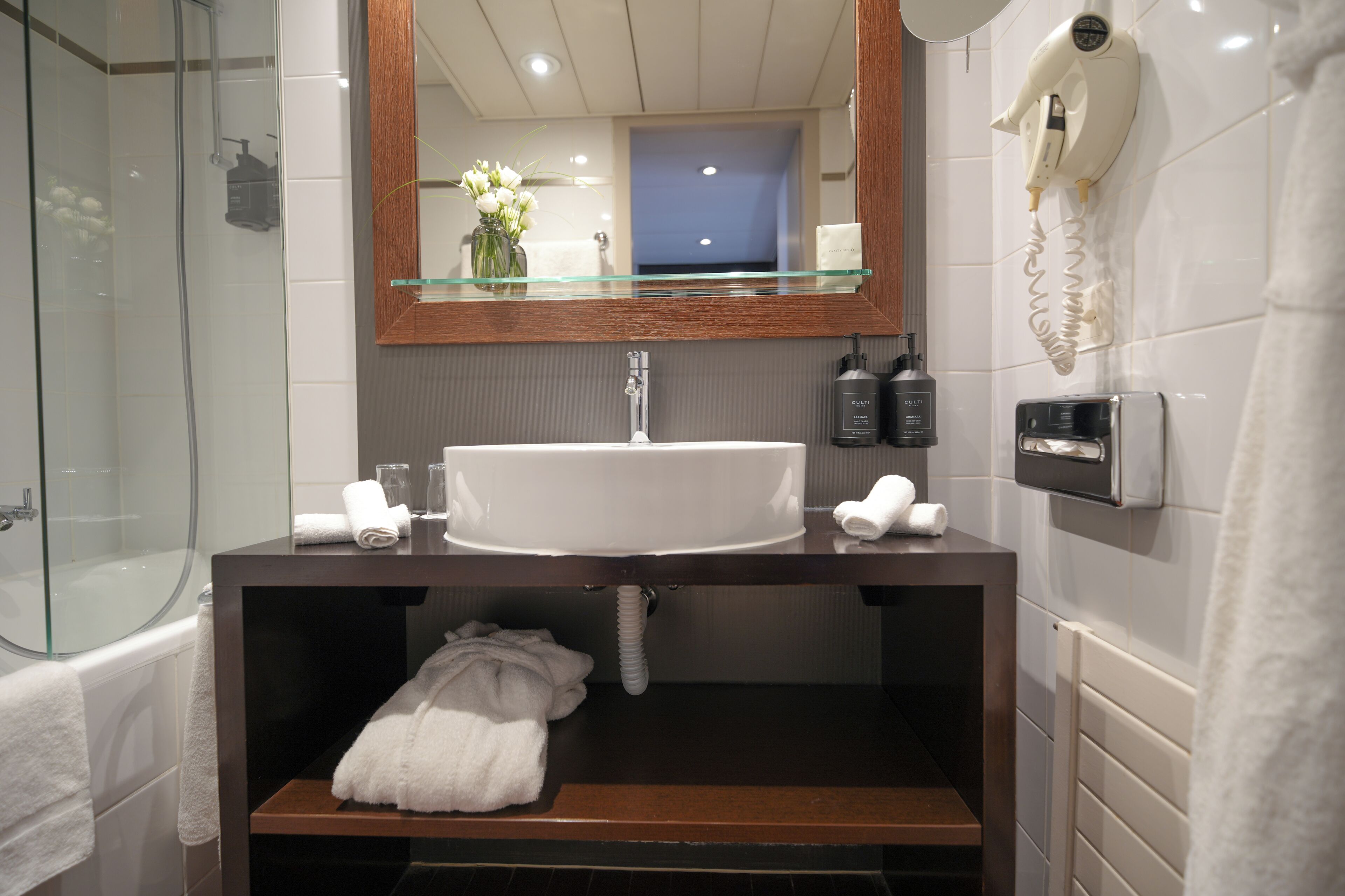 A bathroom with a glass shower enclosure, a white sink, a mirror, and a hairdryer mounted on the wall.