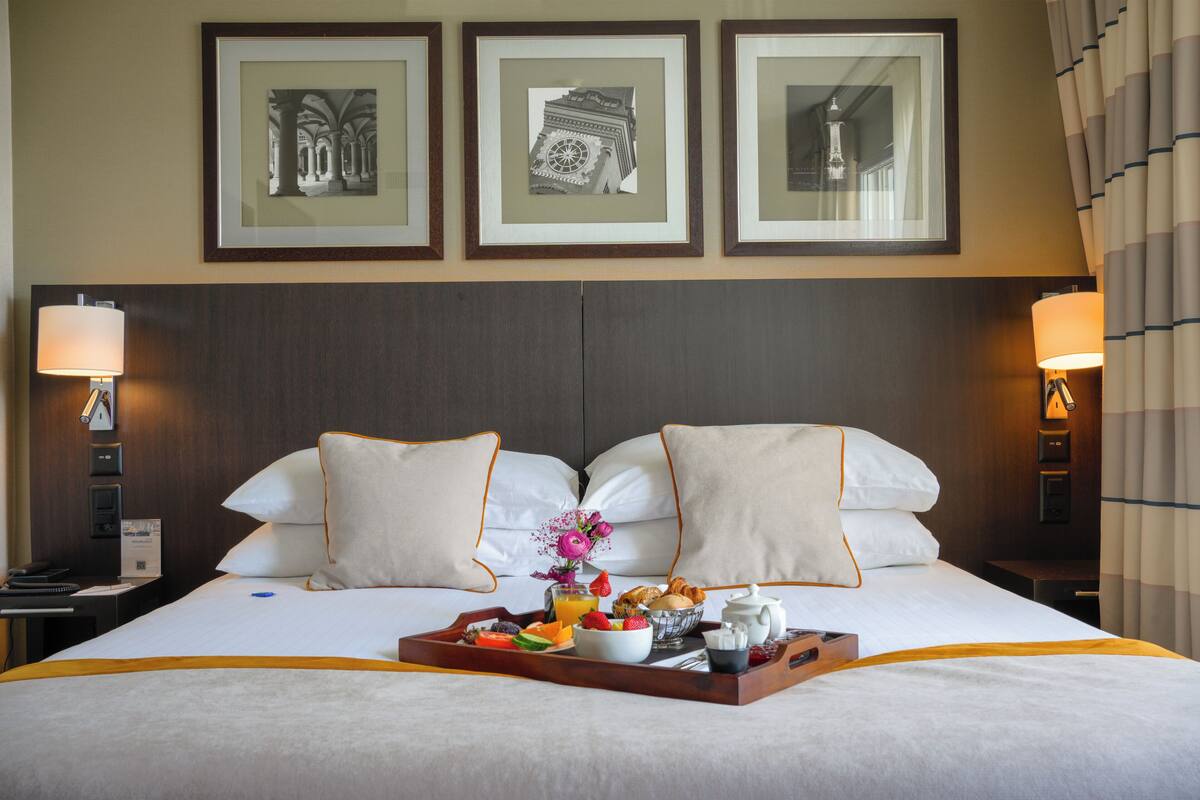 Une chambre d’hôtel avec un lit, des oreillers, un plateau avec le petit-déjeuner, des tableaux encadrés au mur et des lampes.