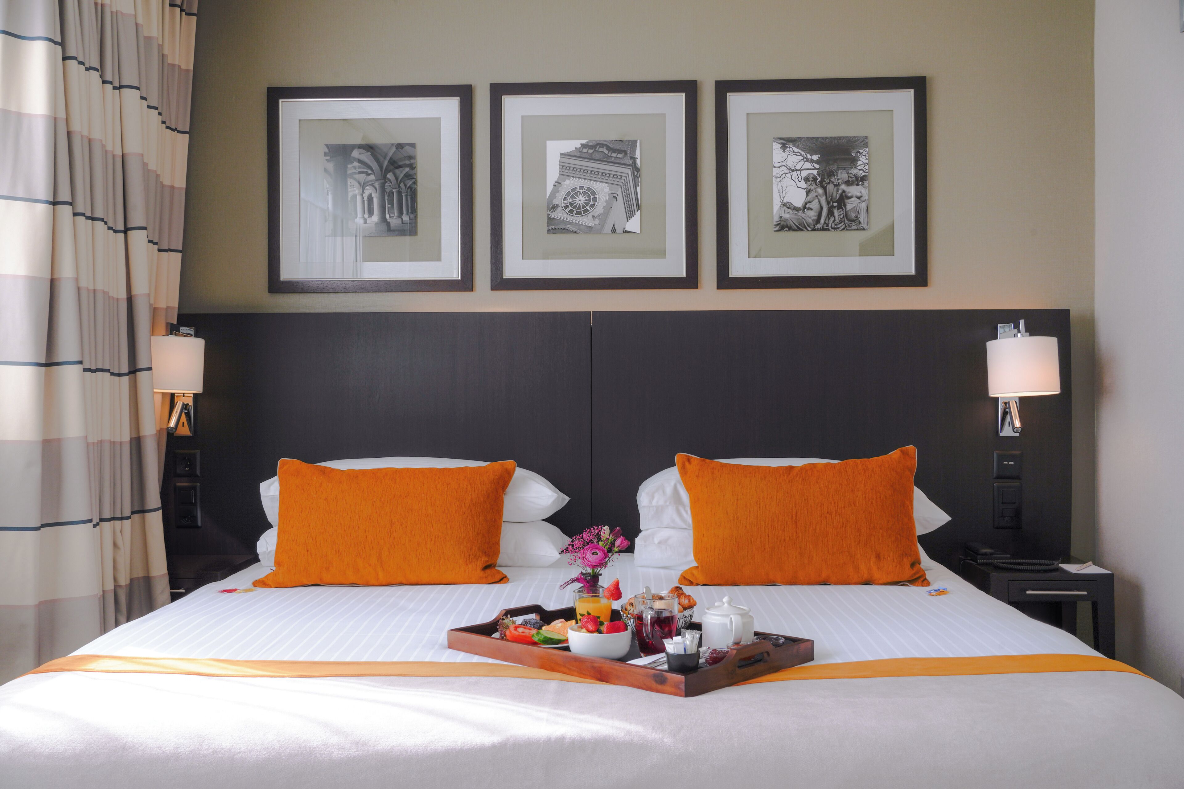 A hotel room with a bed, two pillows, a tray with food and drinks, framed pictures on the wall, and a nightstand with a lamp.
