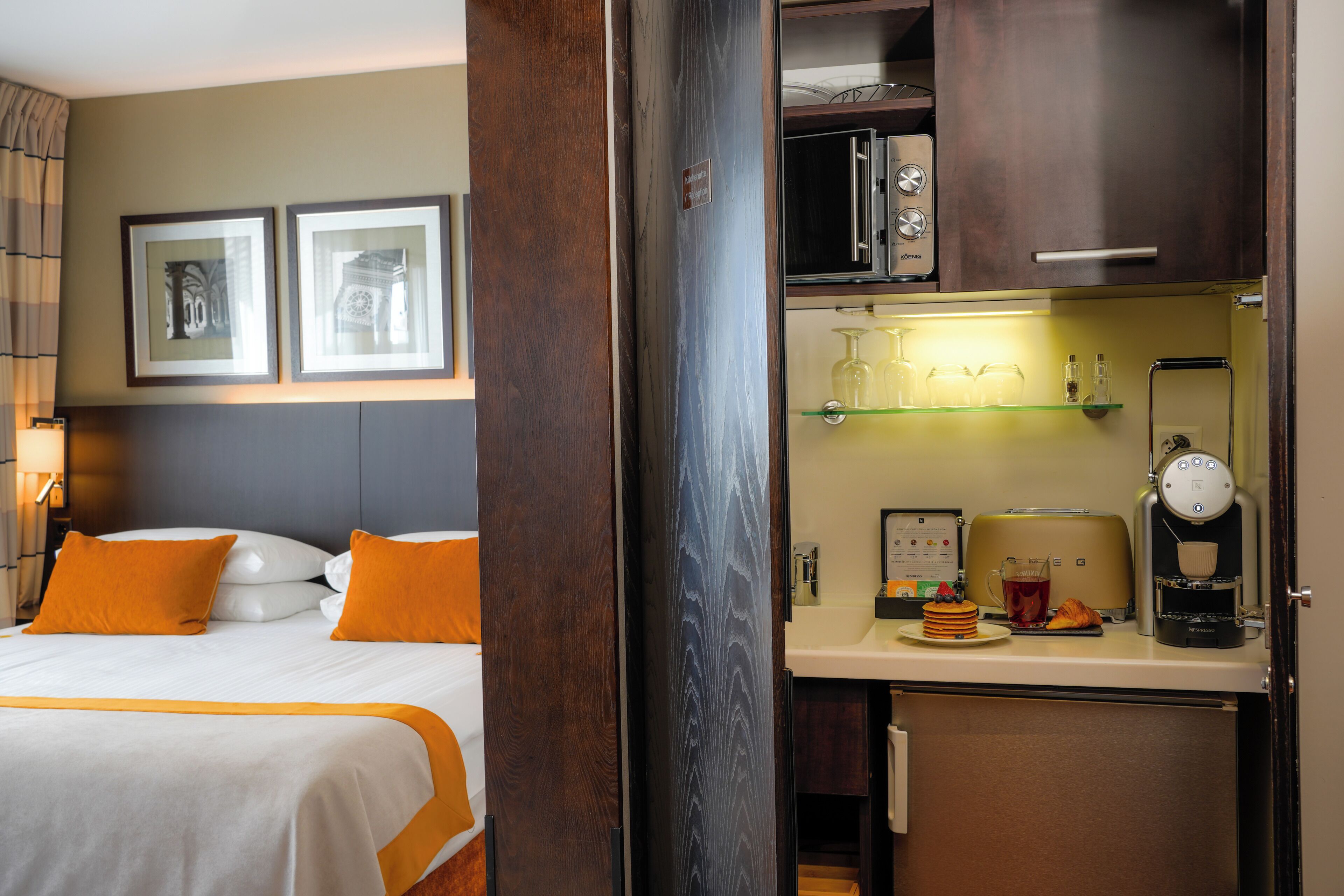 Una camera d'hotel con un letto, un minibar, una macchina del caffè e un ripiano con bicchieri.
