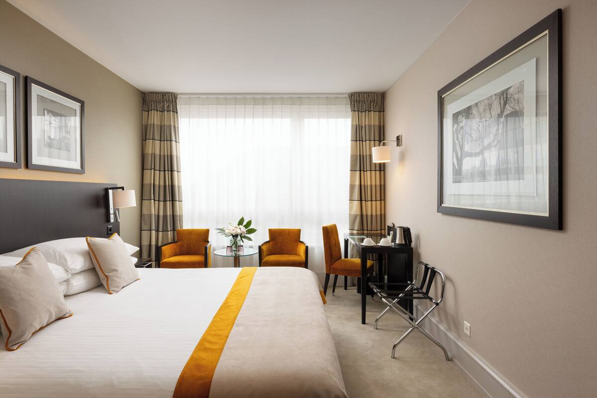 Une chambre d’hôtel avec un grand lit, un bureau avec une chaise, une petite table avec un vase et deux chaises.