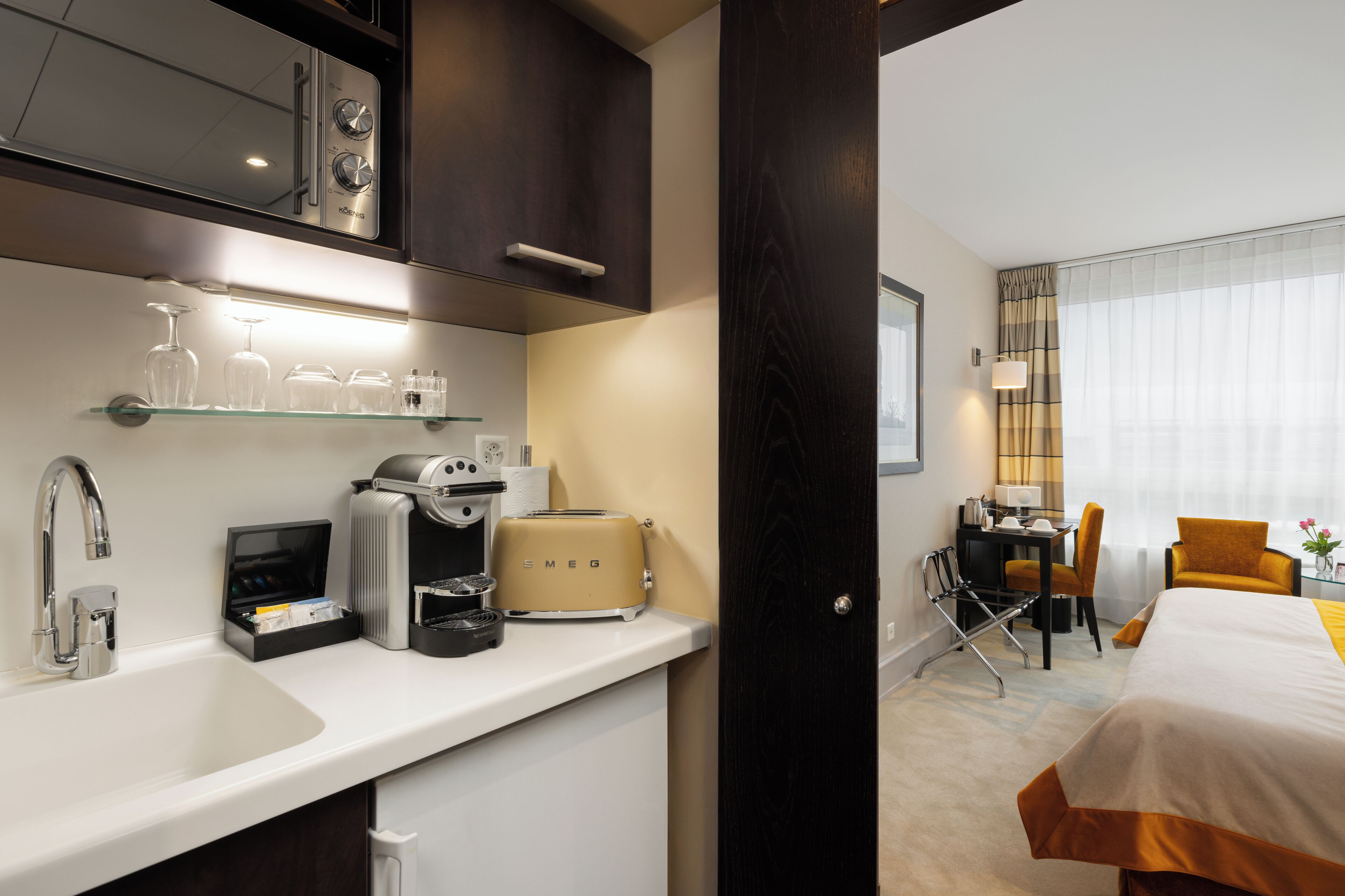 Una habitación de hotel moderna con kitchenette, una cama con detalles naranja, un escritorio con silla y una ventana con cortinas traslúcidas.