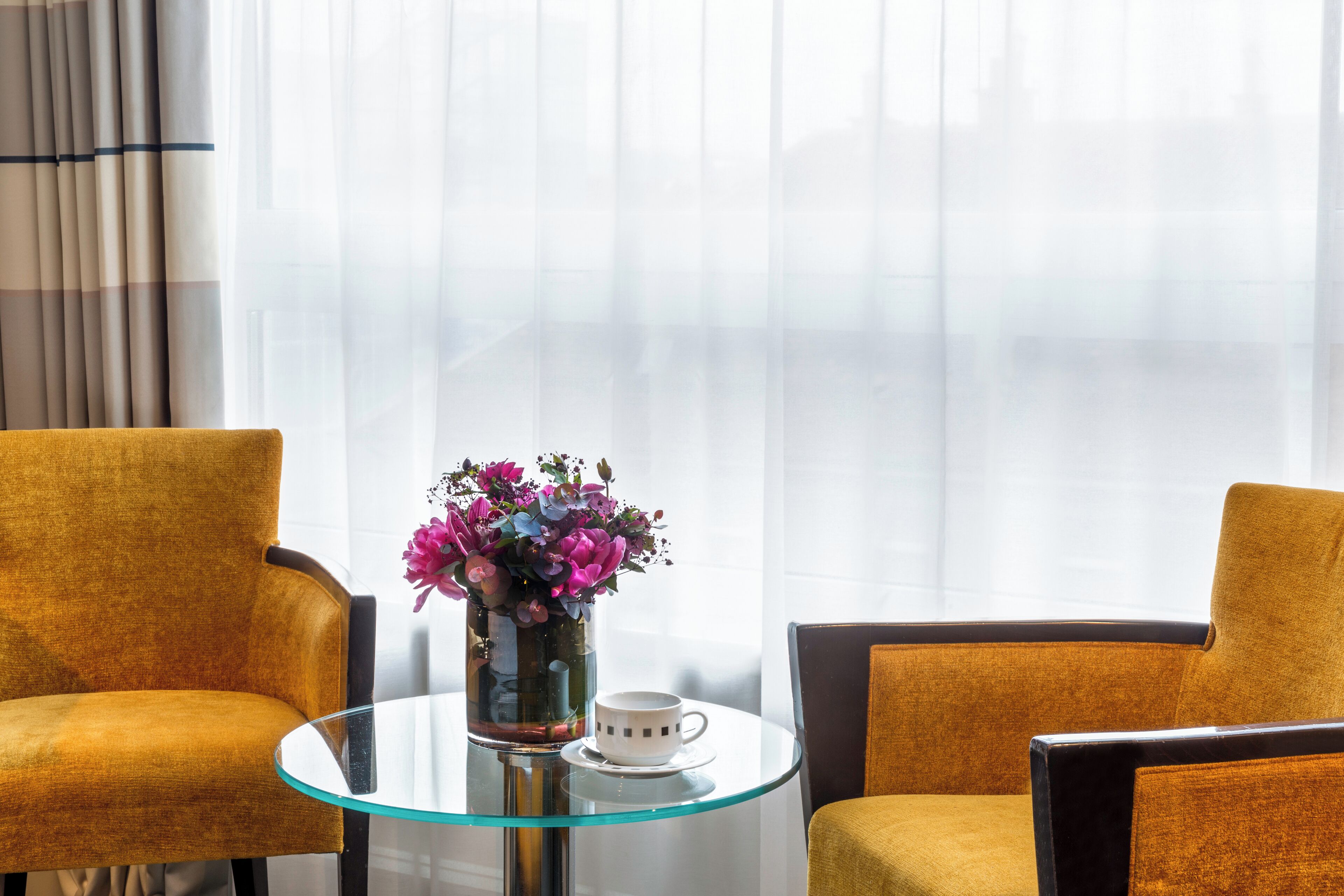 Una habitación con dos sillas color mostaza, una mesa de vidrio con una taza blanca y un florero con flores.