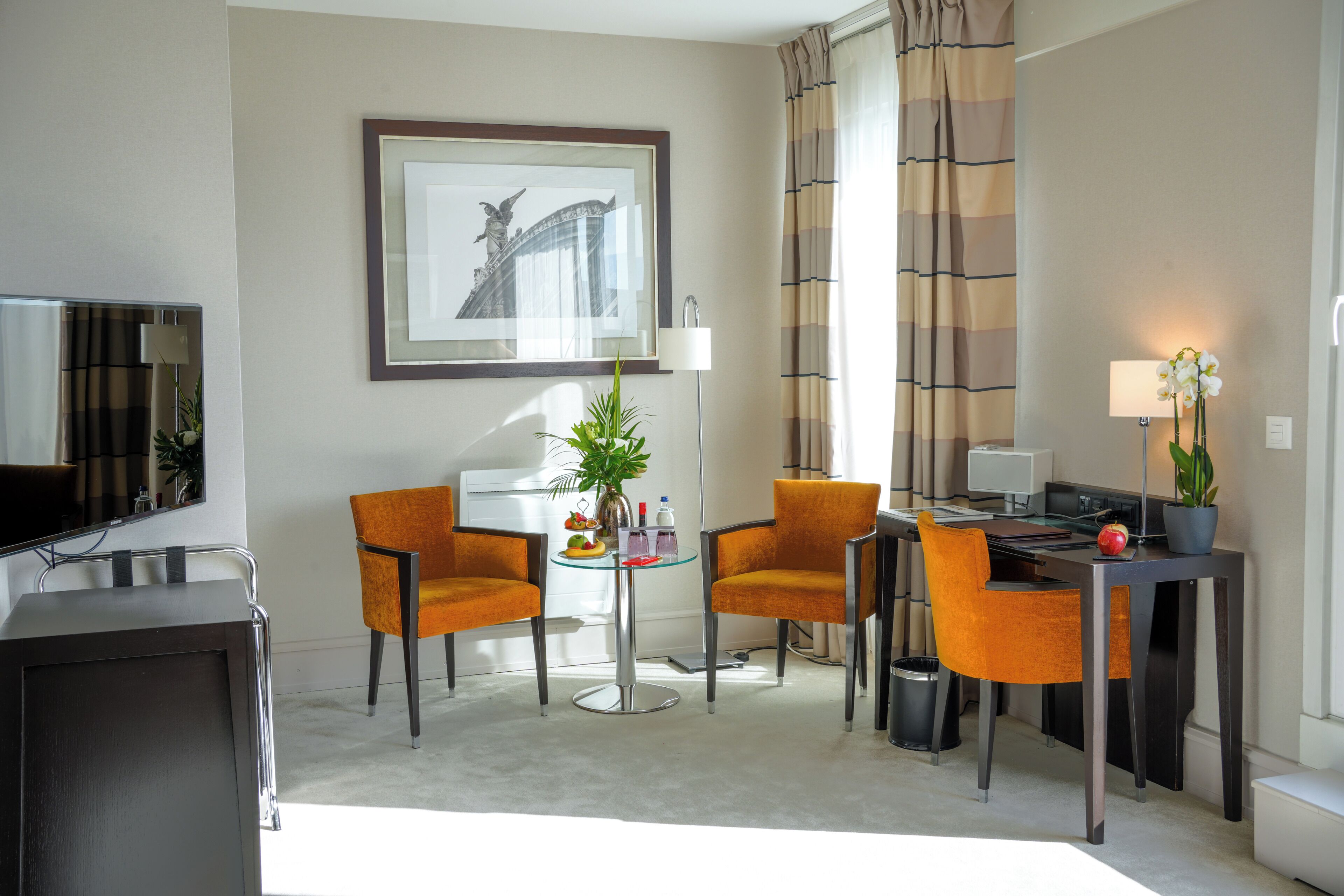 A hotel room with a TV, two orange chairs, a small table with a plant, a framed picture on the wall, and a desk with a lamp and computer.