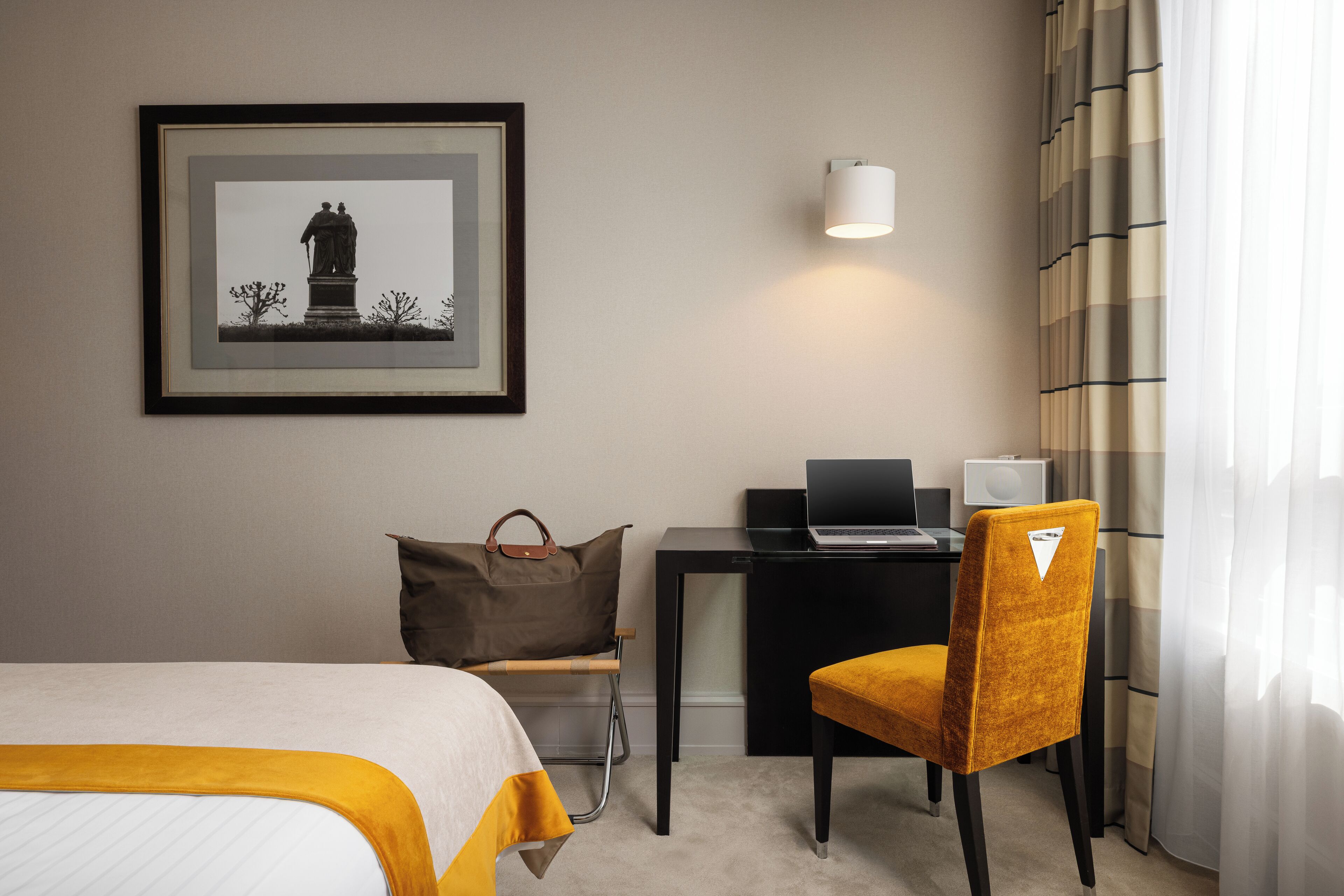 A hotel room with a bed, a desk with a laptop, a chair, a pillow, and a framed picture on the wall.
