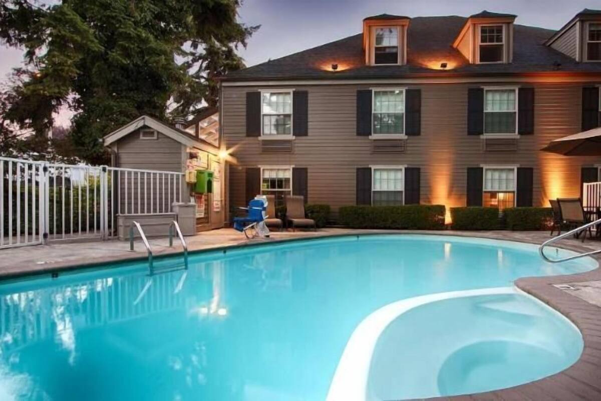 A swimming pool with a wooden deck, a fenced area, and a building with multiple windows and a sloped roof.