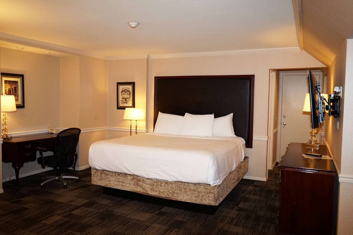 A hotel room with a large bed, a desk with a chair, a desk lamp, a framed picture on the wall, and a door.