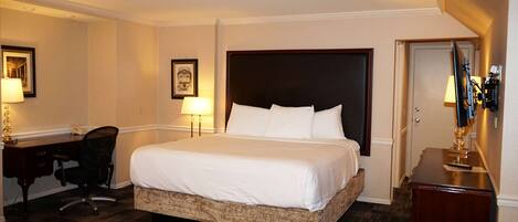 A hotel room with a large bed, a desk with a chair, a desk lamp, a framed picture on the wall, and a door.