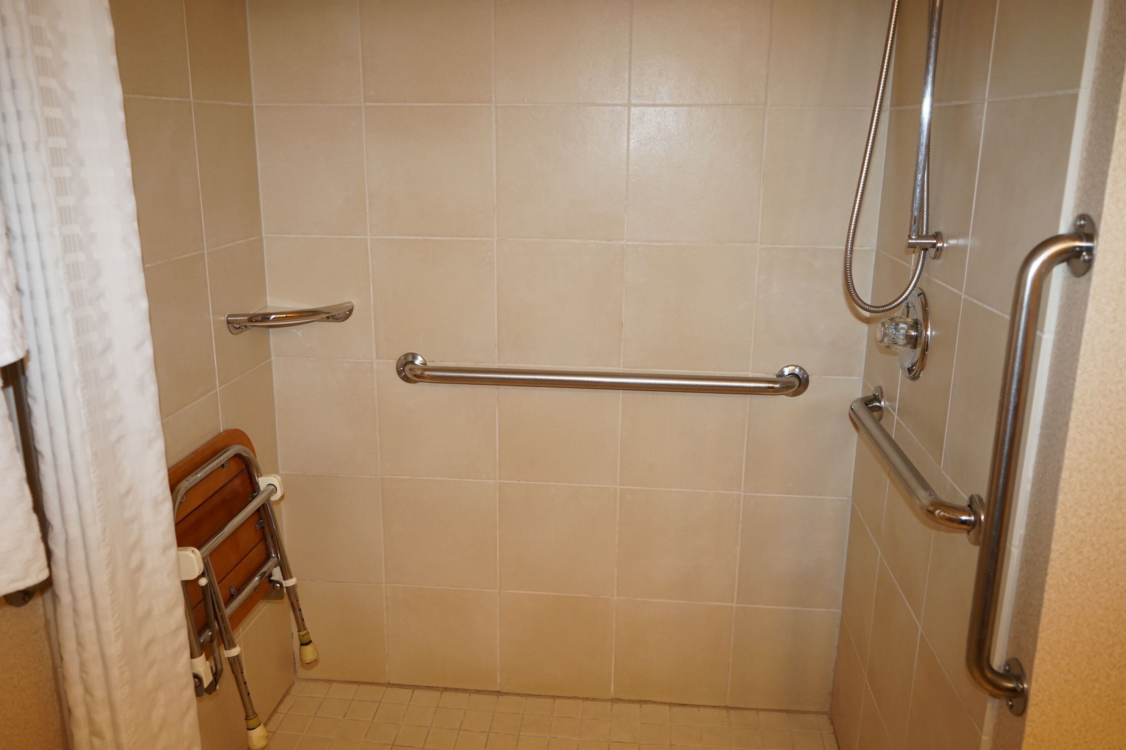A bathroom with beige tiles, a grab bar, a showerhead, and a wooden bench.