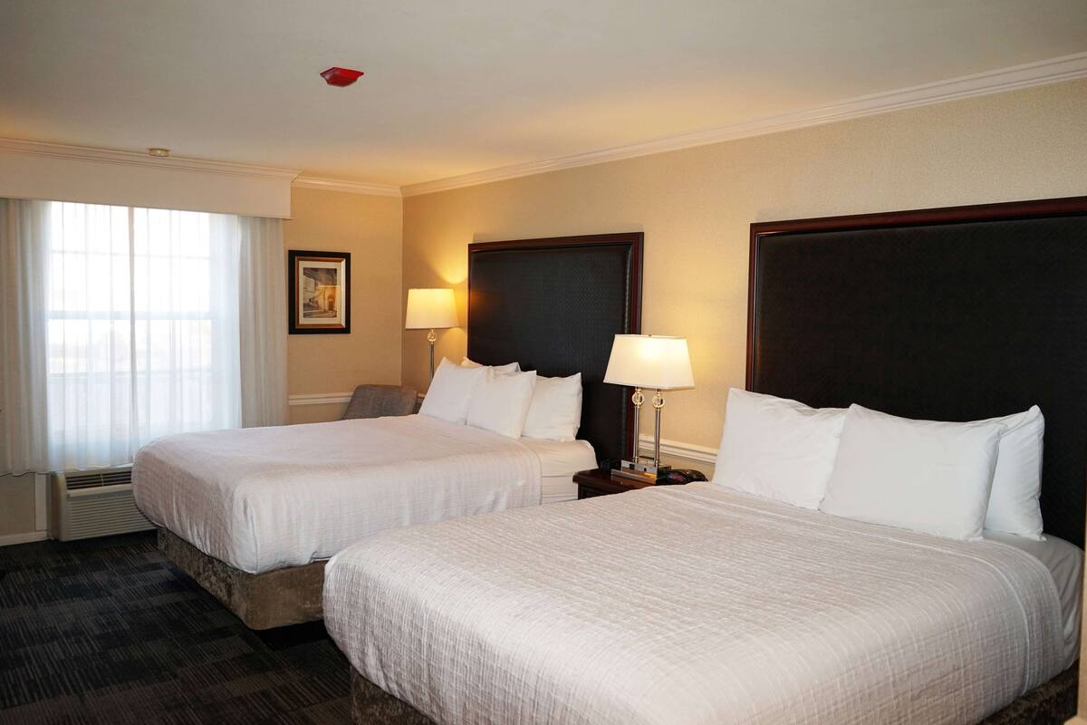 A hotel room with two beds, a large window with curtains, a framed picture on the wall, and two bedside lamps.