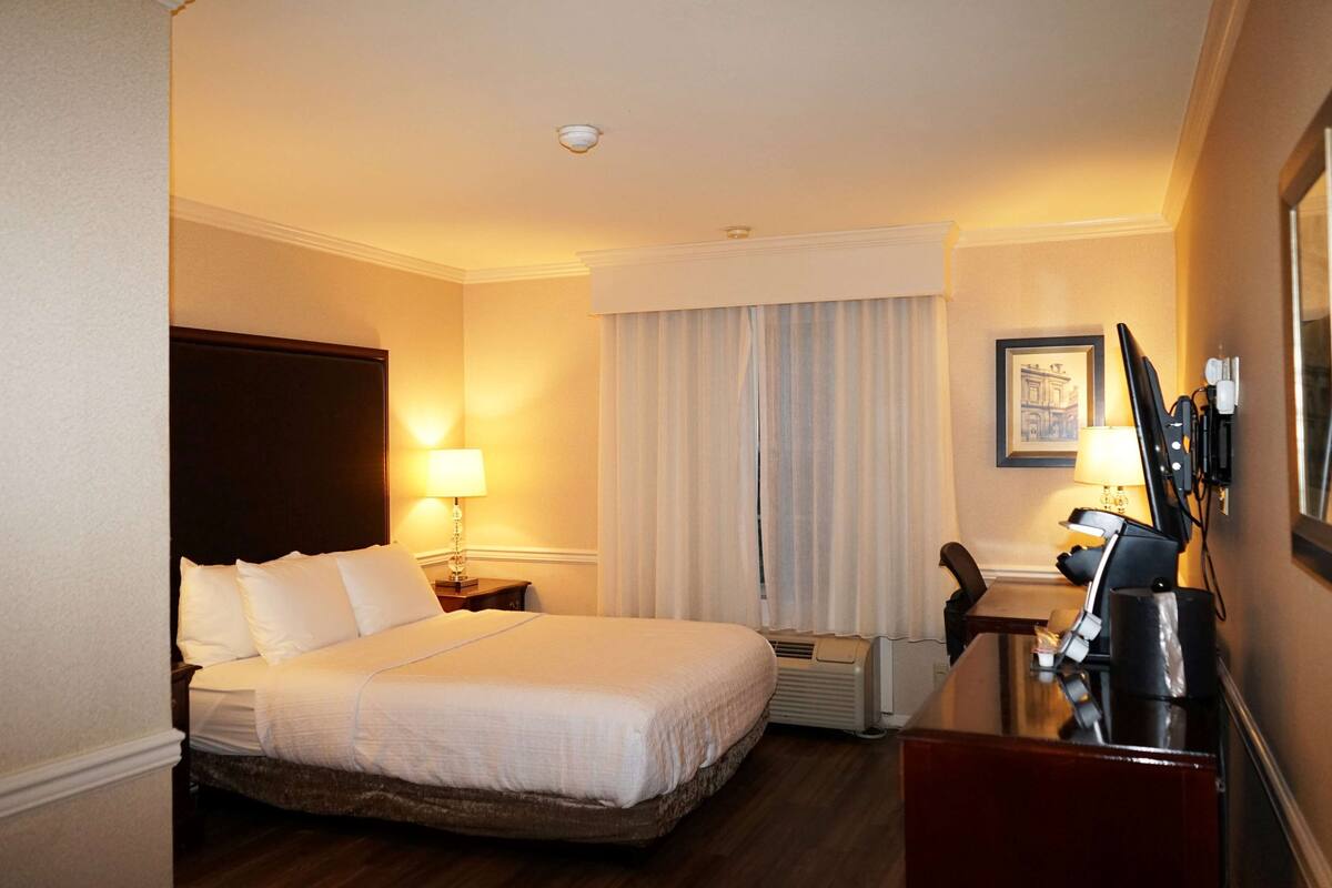 A hotel room with a large bed, a desk with a chair, a lamp, and a framed picture on the wall.