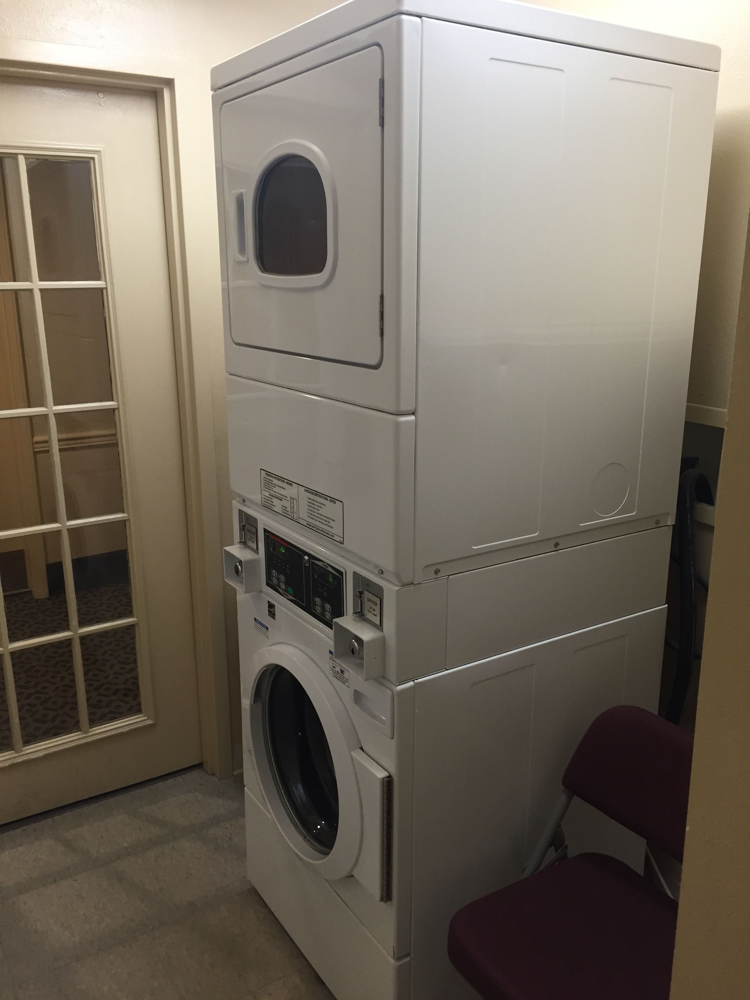 A stacked washer and dryer unit in a laundry room with a door and a chair.