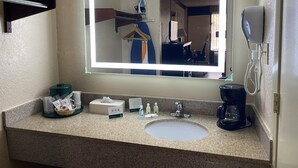 Une salle de bain d’hôtel avec un comptoir en granit, un lavabo blanc, un miroir avec éclairage, un sèche-cheveux et une machine à café.