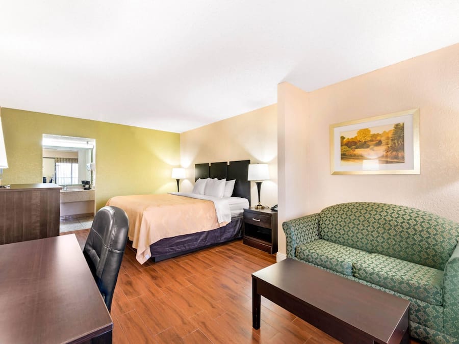 A hotel room with a bed, a green patterned sofa, a wooden table, a desk, and a painting on the wall.