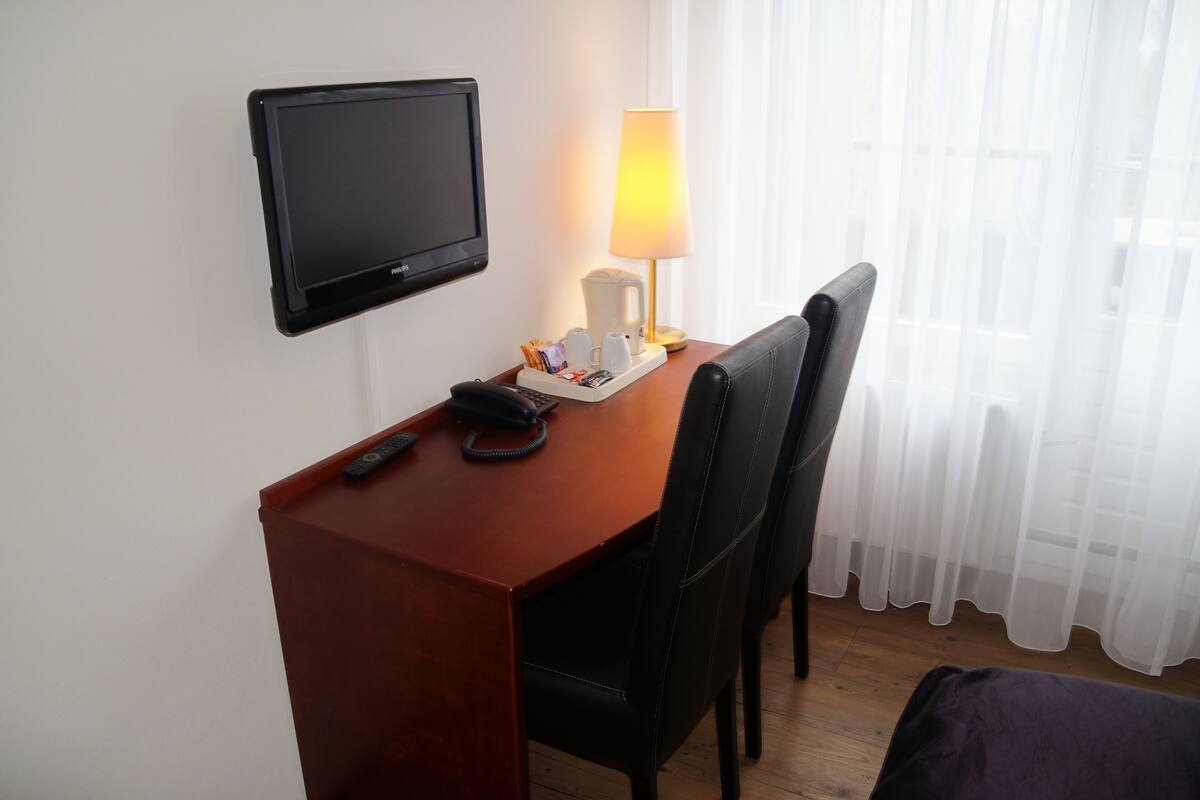 A hotel room with a wooden desk, a black chair, a television, a lamp, and a coffee maker.
