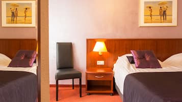 Habitación de hotel con dos camas, una mesita de noche con lámpara, una silla y dos cuadros en la pared.