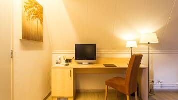 A hotel room with a wooden desk, a computer monitor, a chair, a lamp, and a painting on the wall.
