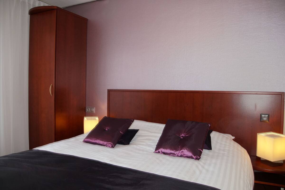 A hotel room with a large wooden wardrobe, a bed with white bedding and purple pillows, and bedside lamps.